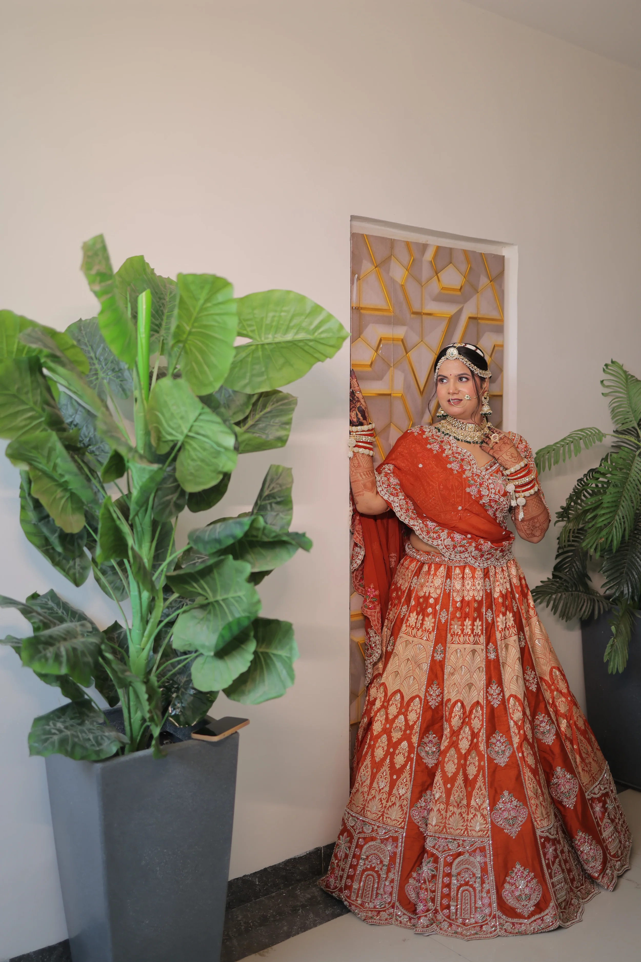 A woman wearing a red and gold traditional Indian bridal saree with jewelry, including earrings, necklace, and headpiece, standing in a decorative doorway. Large green plants are visible on either side of her, one in a gray pot and the other plants w