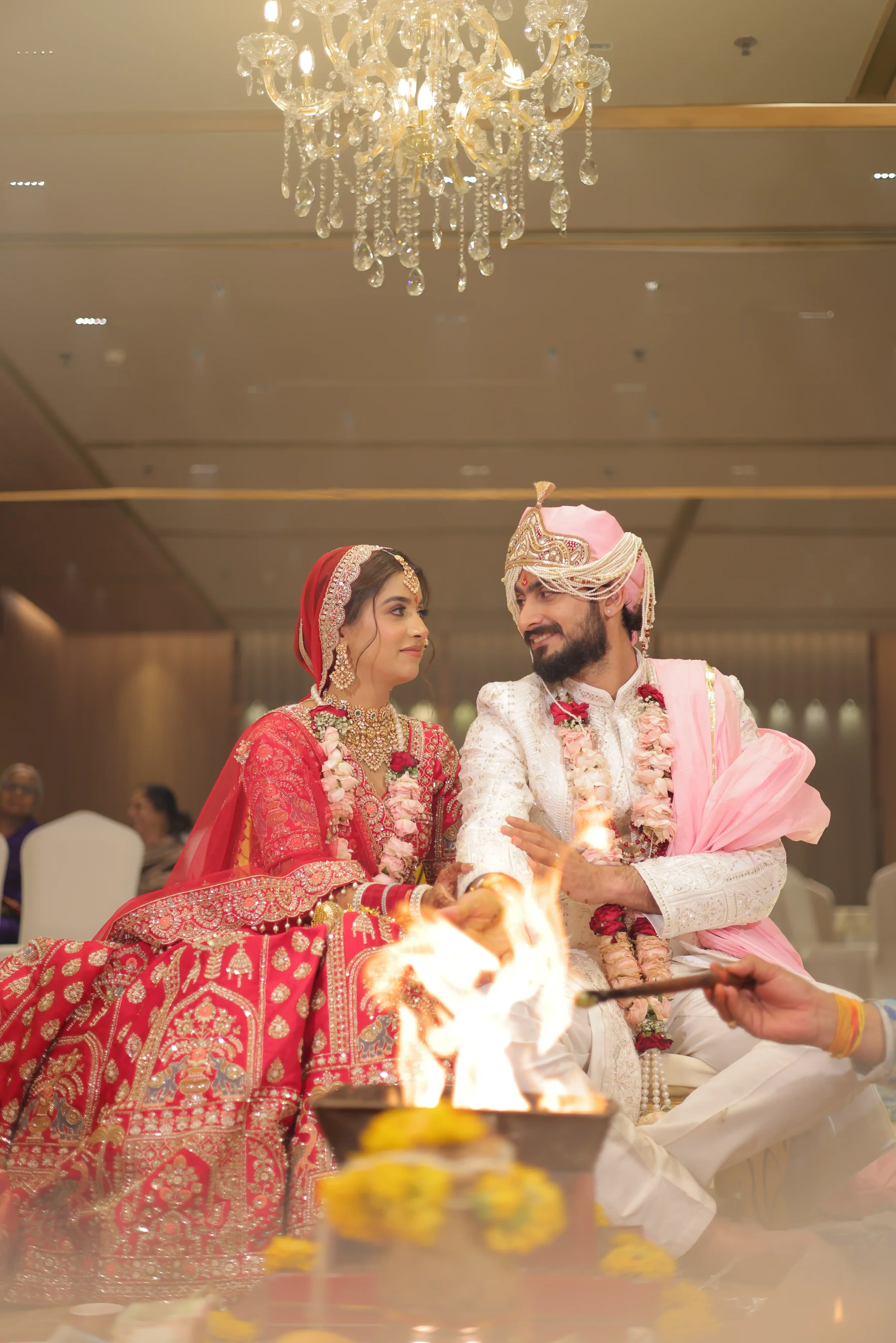 A bride and groom dressed in traditional Indian wedding attire participating in a fire ritual during their wedding ceremony, with a chandelier hanging above and other guests seated in the background.