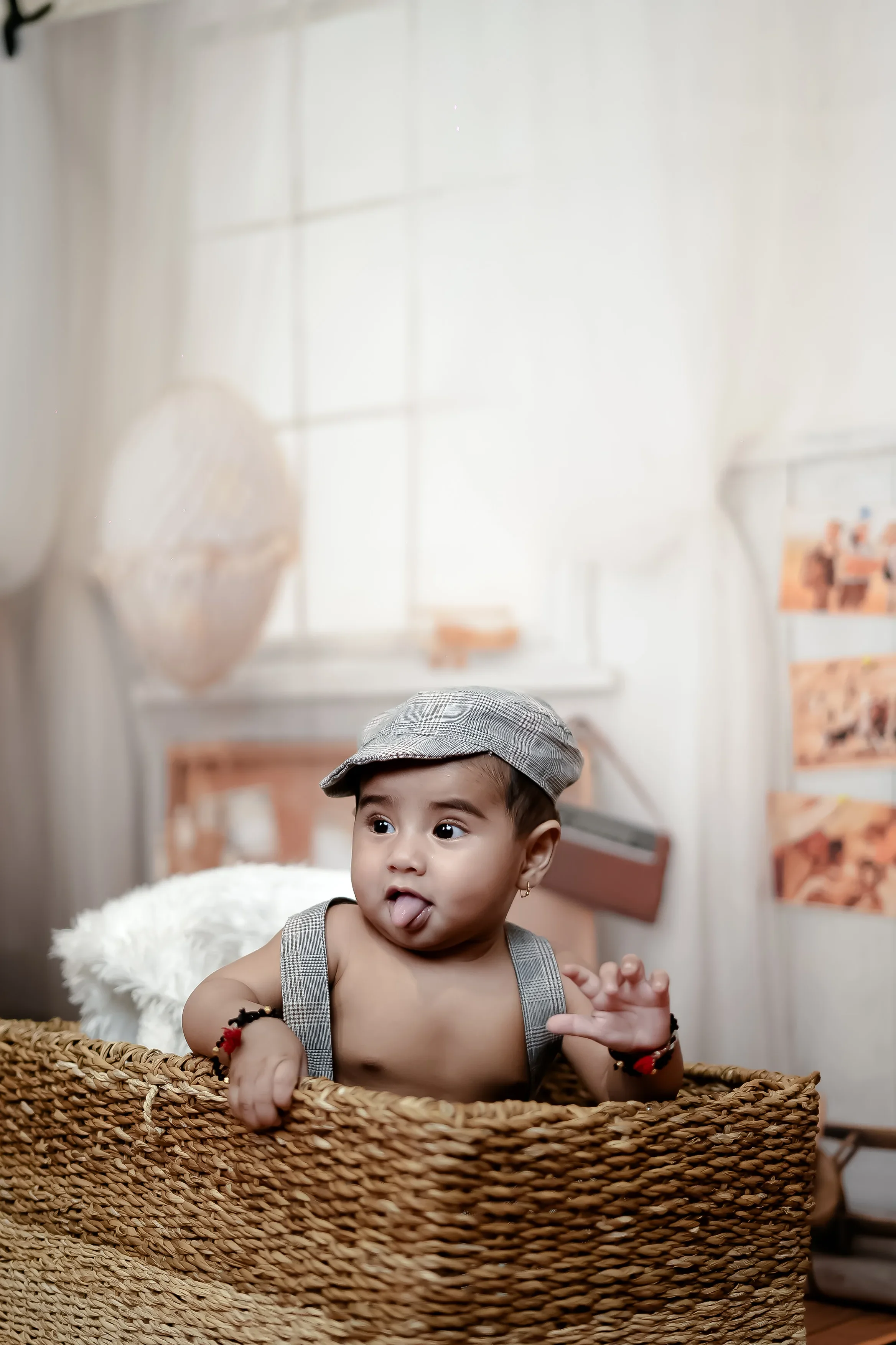 A young child with dark hair, wearing a plaid cap and a sleeveless checkered outfit, sitting in a woven basket indoors with a blurred background of posters and a window. The child has a playful expression with their tongue sticking out and is holding