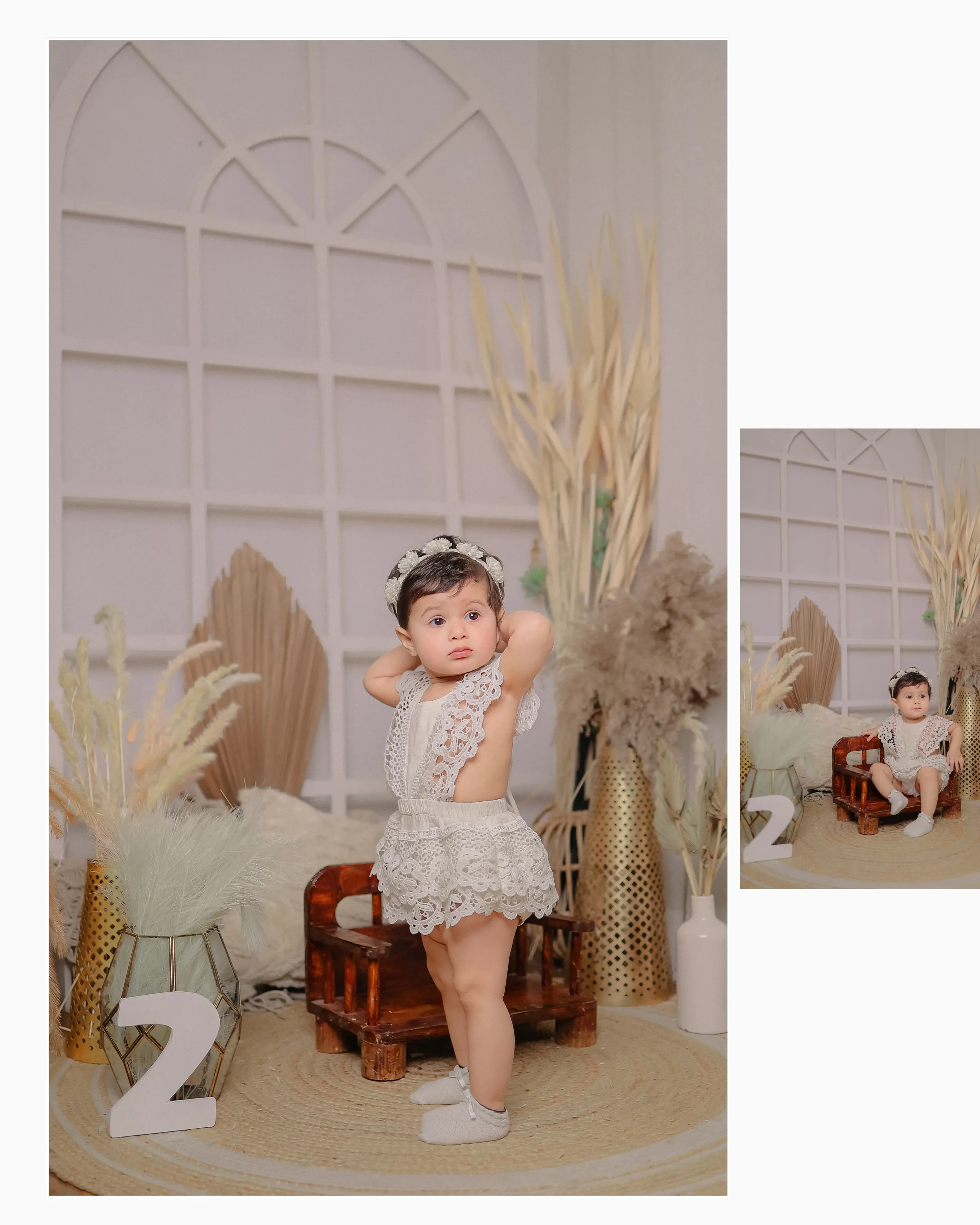 A young girl with short hair, wearing a white lace dress and white shoes, stands on a beige rug in front of a decorative backdrop with large pampas grass and stylish vases, in a setting marked by the number 2.