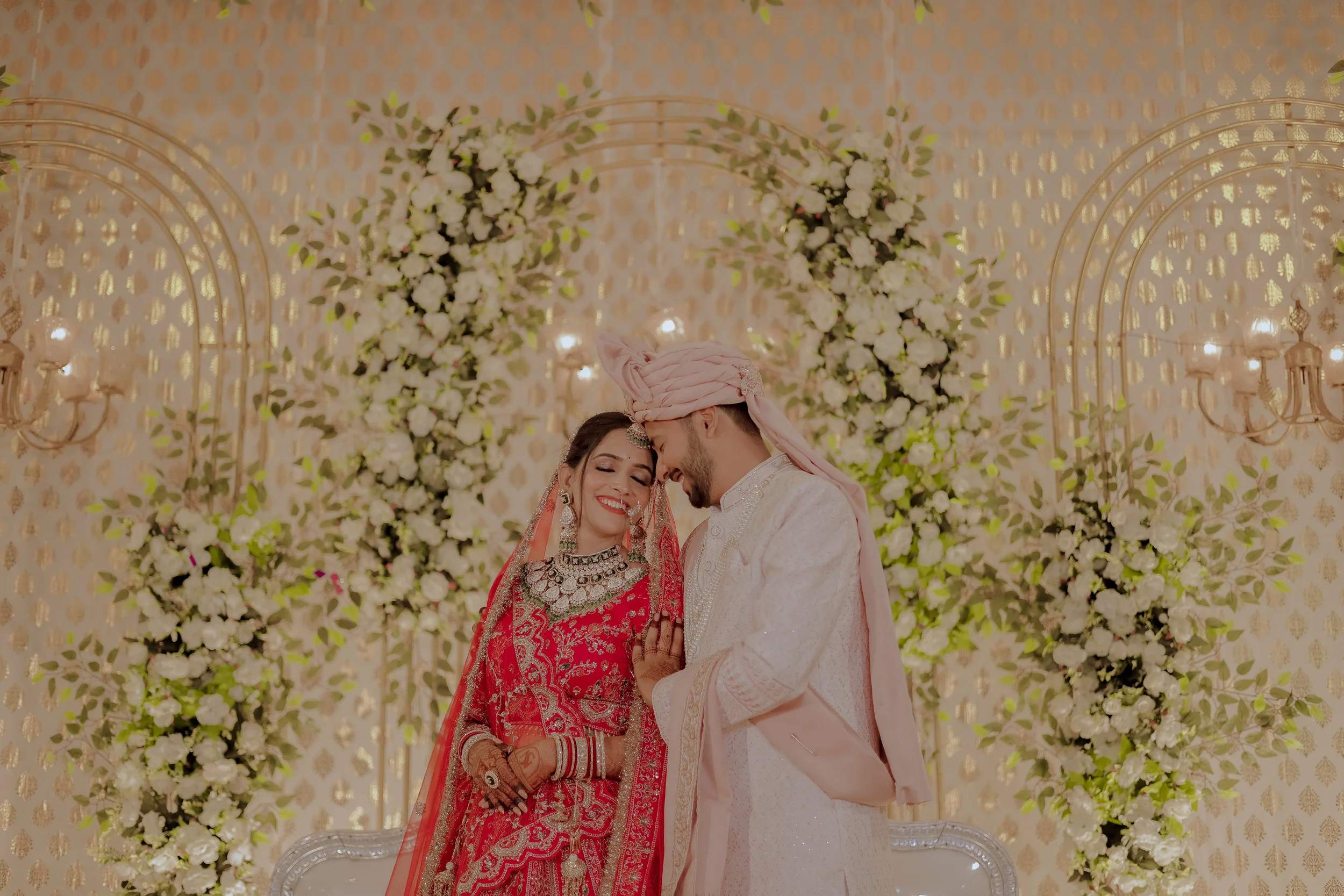 A bride and groom celebrating their wedding, standing close with foreheads touching, surrounded by white flowers and elegant gold decor.