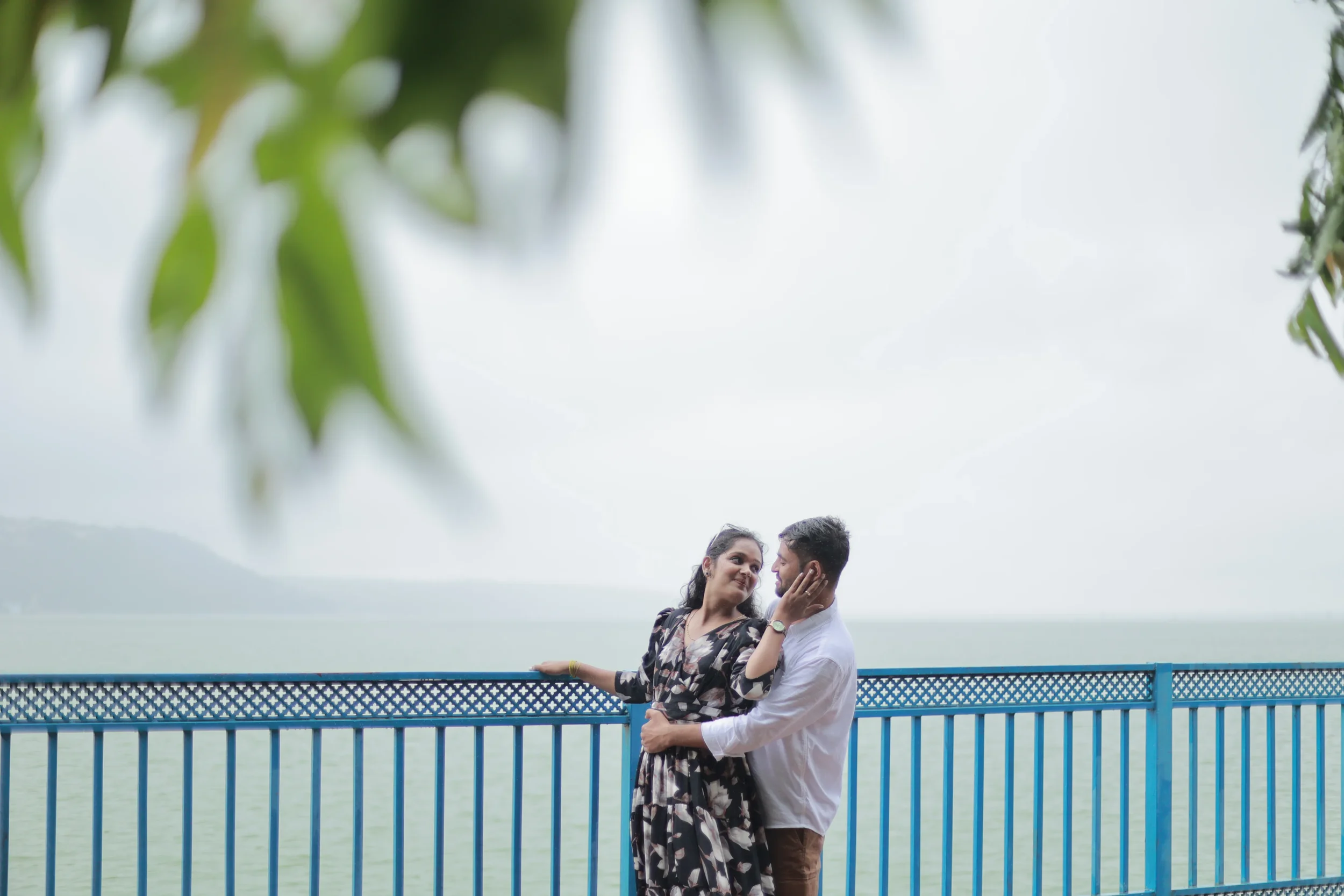 A couple stands close on a blue bridge near water, with the woman touching the man's face, both smiling, in a scenic outdoor setting with trees and cloudy sky.