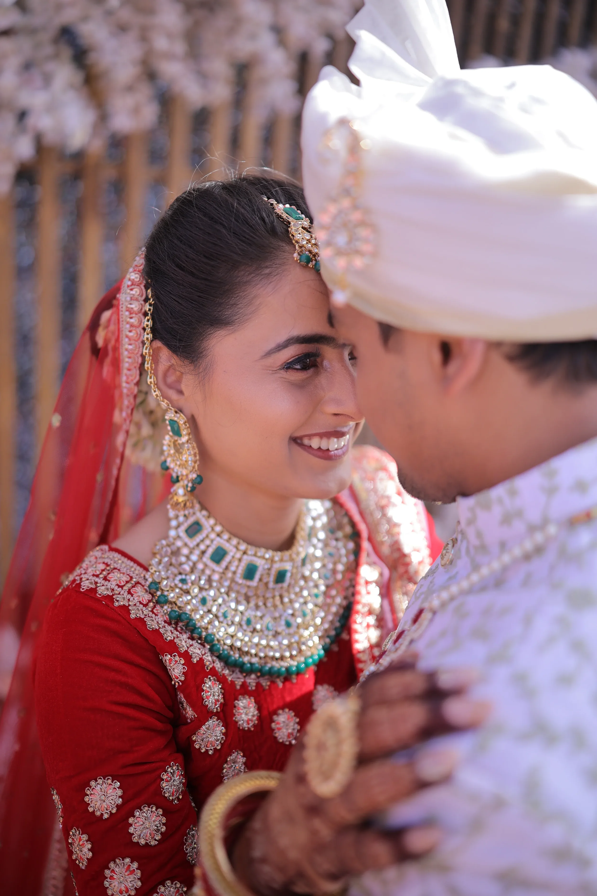 Close-up of a smiling bride in traditional Indian wedding attire and jewelry, touching foreheads with a groom wearing a white turban and outfit, during a wedding ceremony.