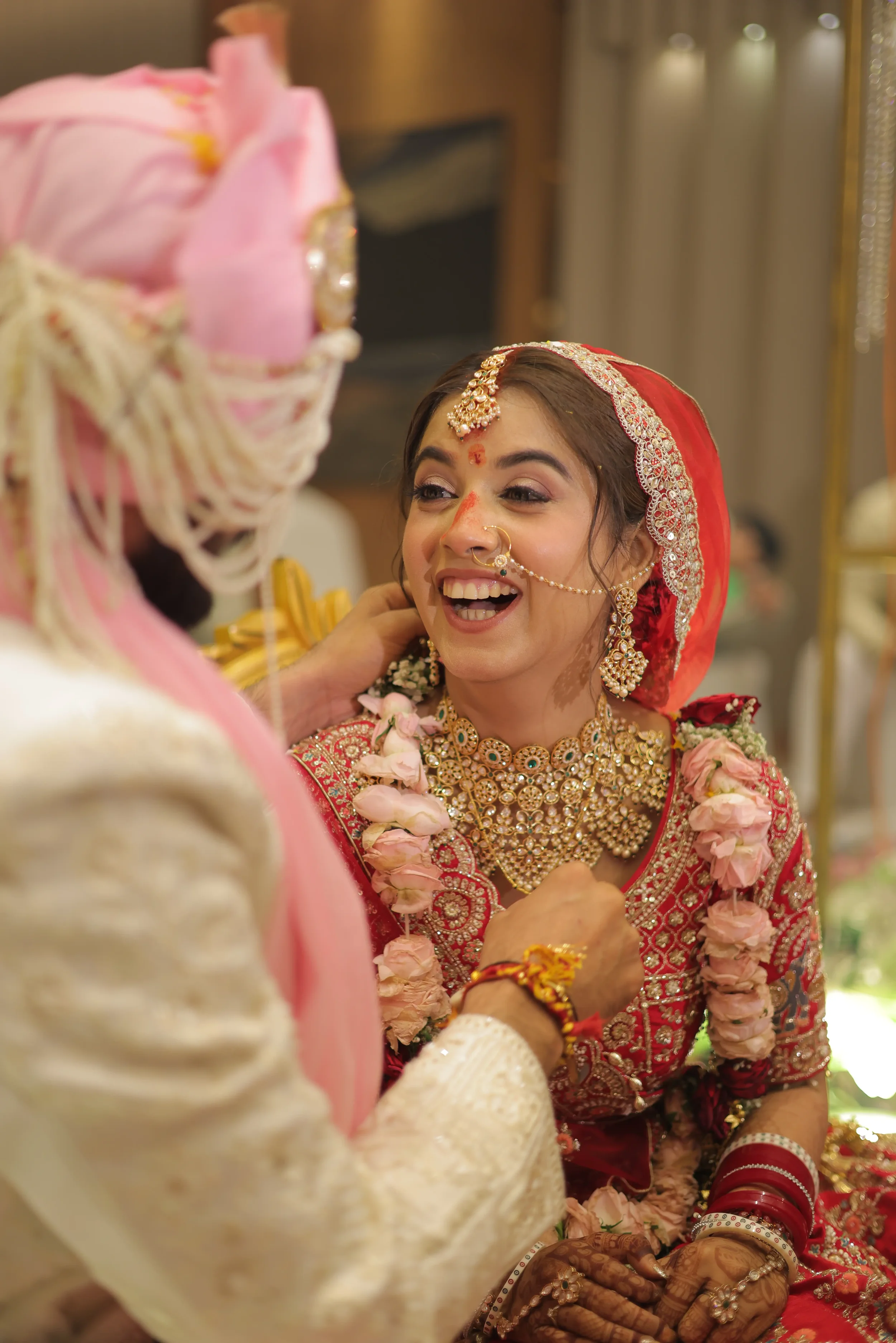 A bride dressed in traditional red and gold Indian wedding attire, wearing jewelry, and decorated with flower garlands, smiling during a wedding ceremony. A groom in traditional attire is visible, gently touching her face.