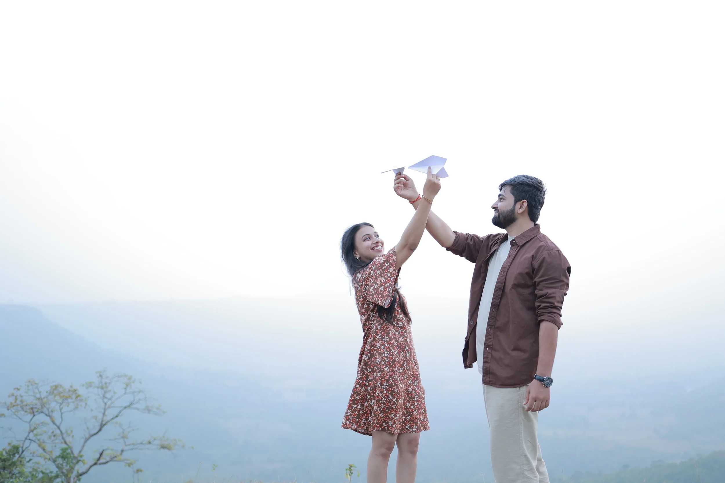 A woman and a man outdoors on a cloudy day, holding paper airplanes and smiling at each other.