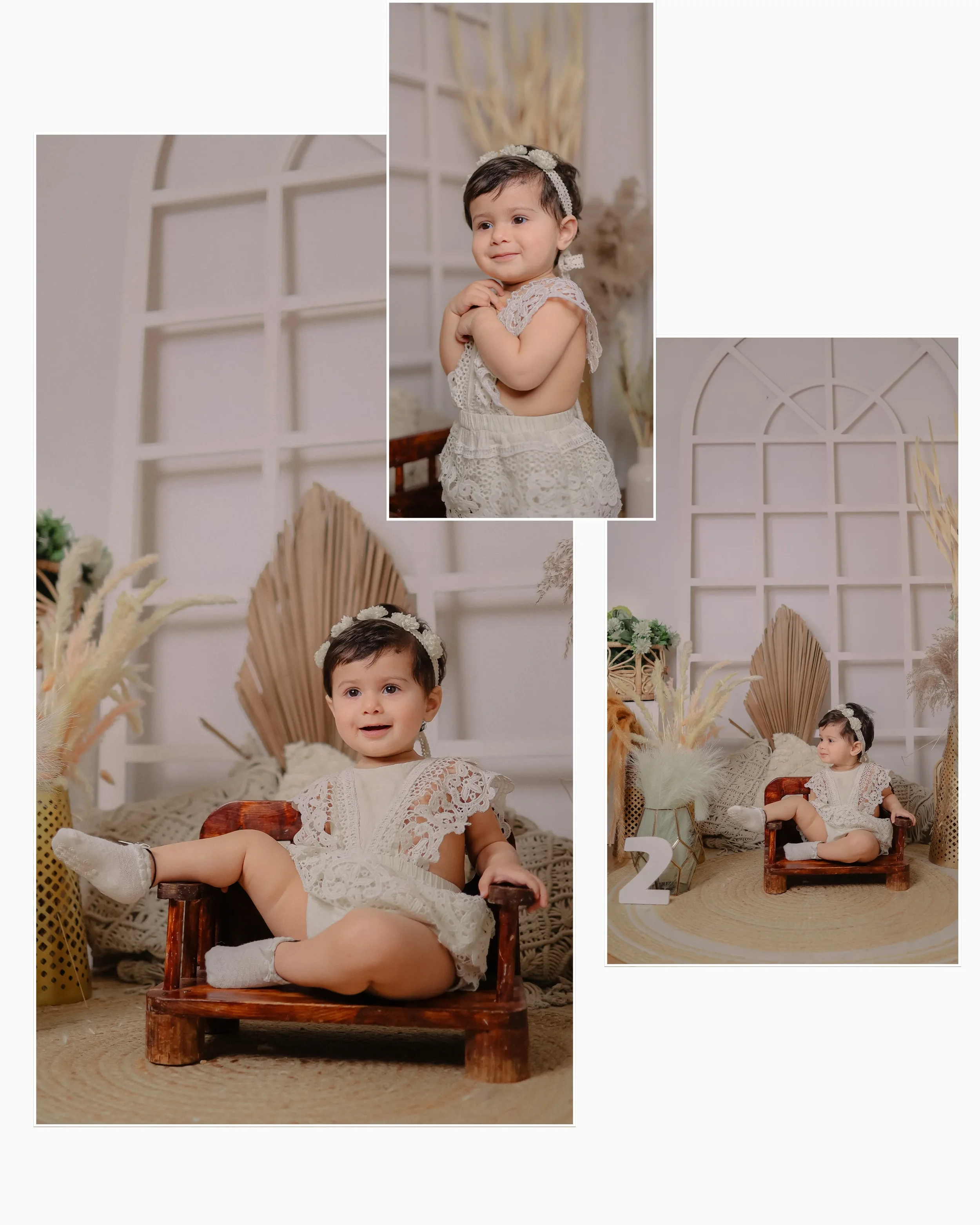A little girl with short dark hair, wearing a white lace dress, sits on a small wooden chair with her legs crossed. The background features white decorative panels, dried plants, and cushions, creating a cozy, neutral-toned setting.