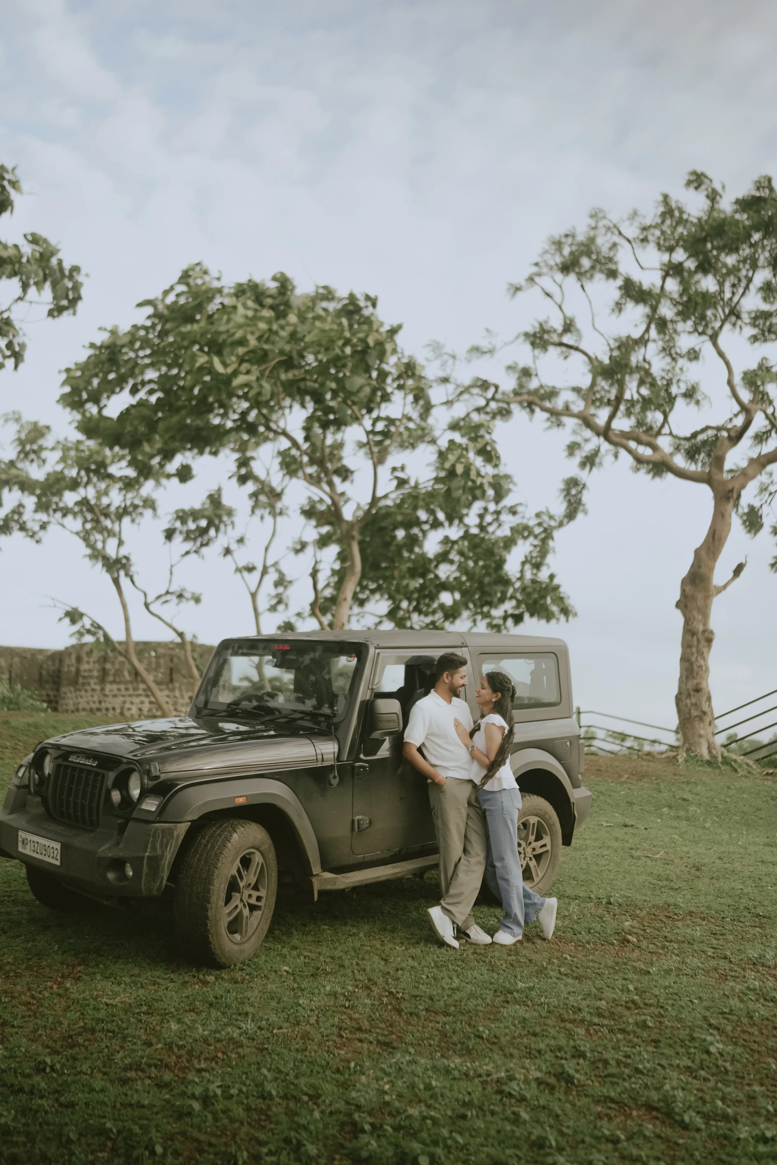 A couple stands close together, smiling, with the man leaning against a black SUV and the woman standing nearby. They are outdoors on a grassy area with large trees and a cloudy sky in the background.