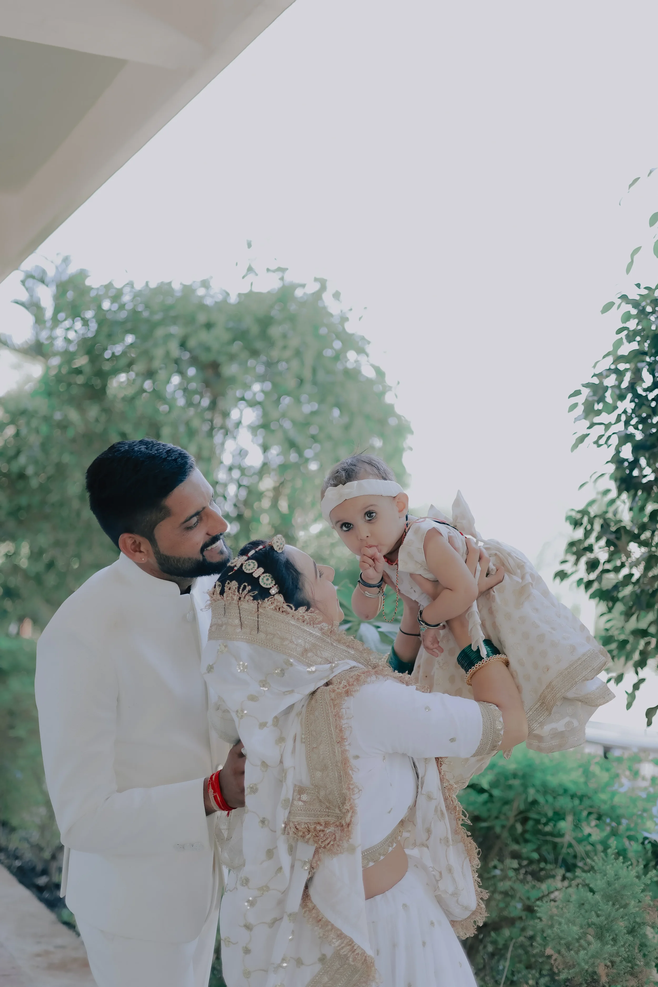 A man dressed in traditional white Indian attire holding a young girl dressed in traditional cream and gold attire. The girl is also wearing a headband and is touching her lips with her finger. They are outdoors with green trees and foliage in the ba