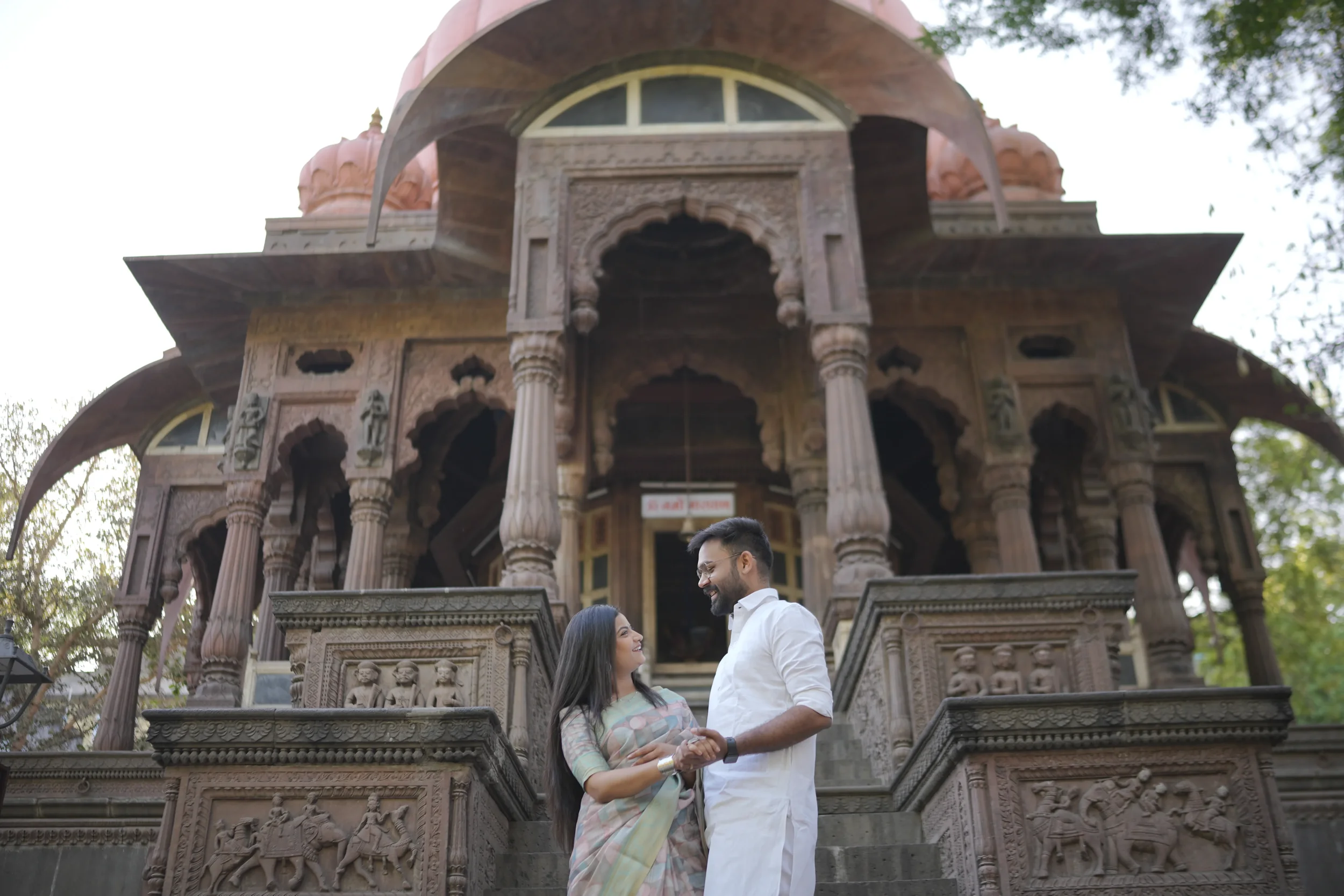 A man and woman stand on steps in front of an ornate, historic building with detailed carvings and multiple arches, shaking hands and smiling at each other.