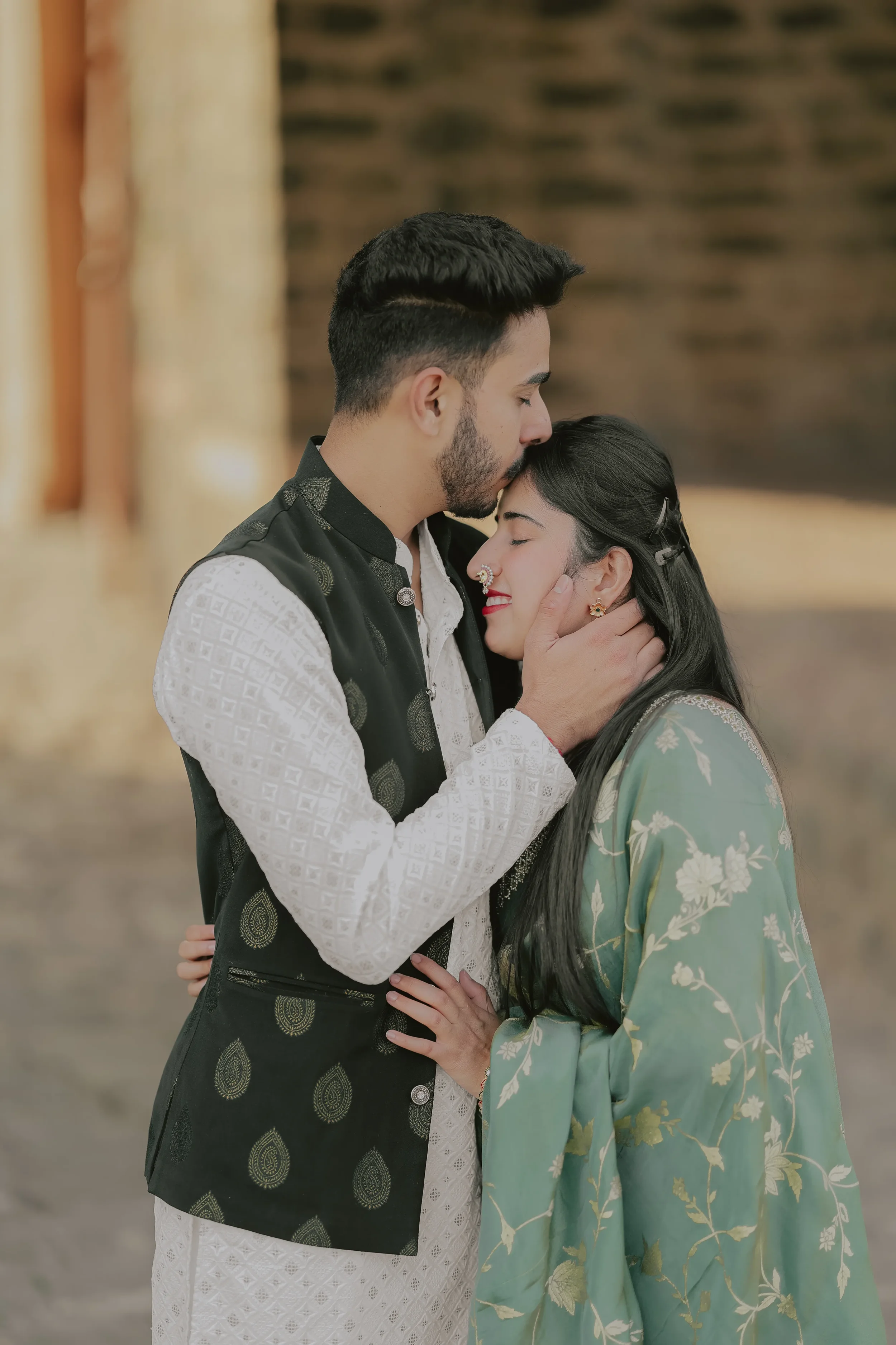 A man and woman sharing a tender moment with the man holding the woman's face and the woman closing her eyes and smiling, wearing traditional Indian clothing, indoors with a blurred background.