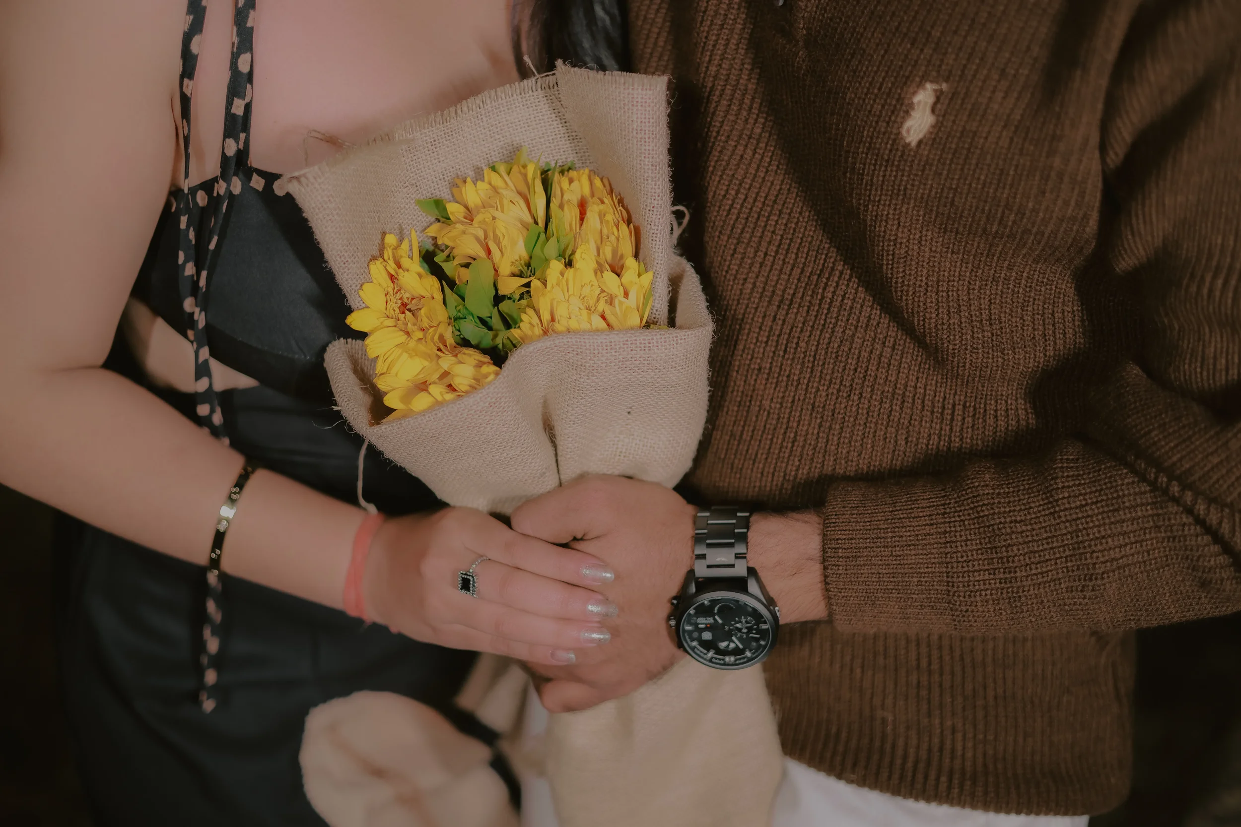 Two people holding a bouquet of yellow flowers together, one wearing a black and pink dress and jewelry, the other wearing a brown sweater and watch, standing close with their hands intertwined.