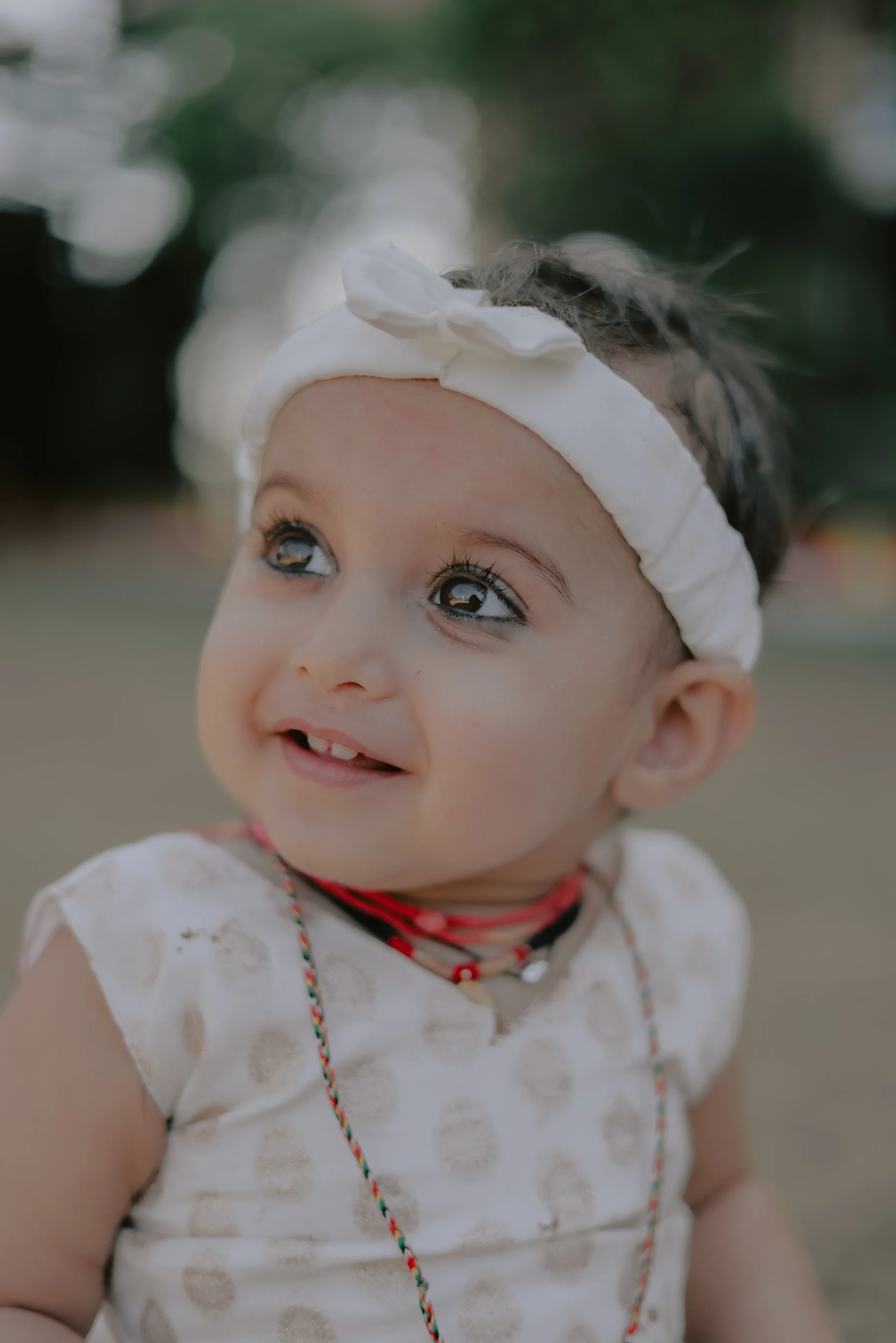 Close-up of a young girl wearing a white headband with a bow, smiling and looking up, outdoors with blurred green background.