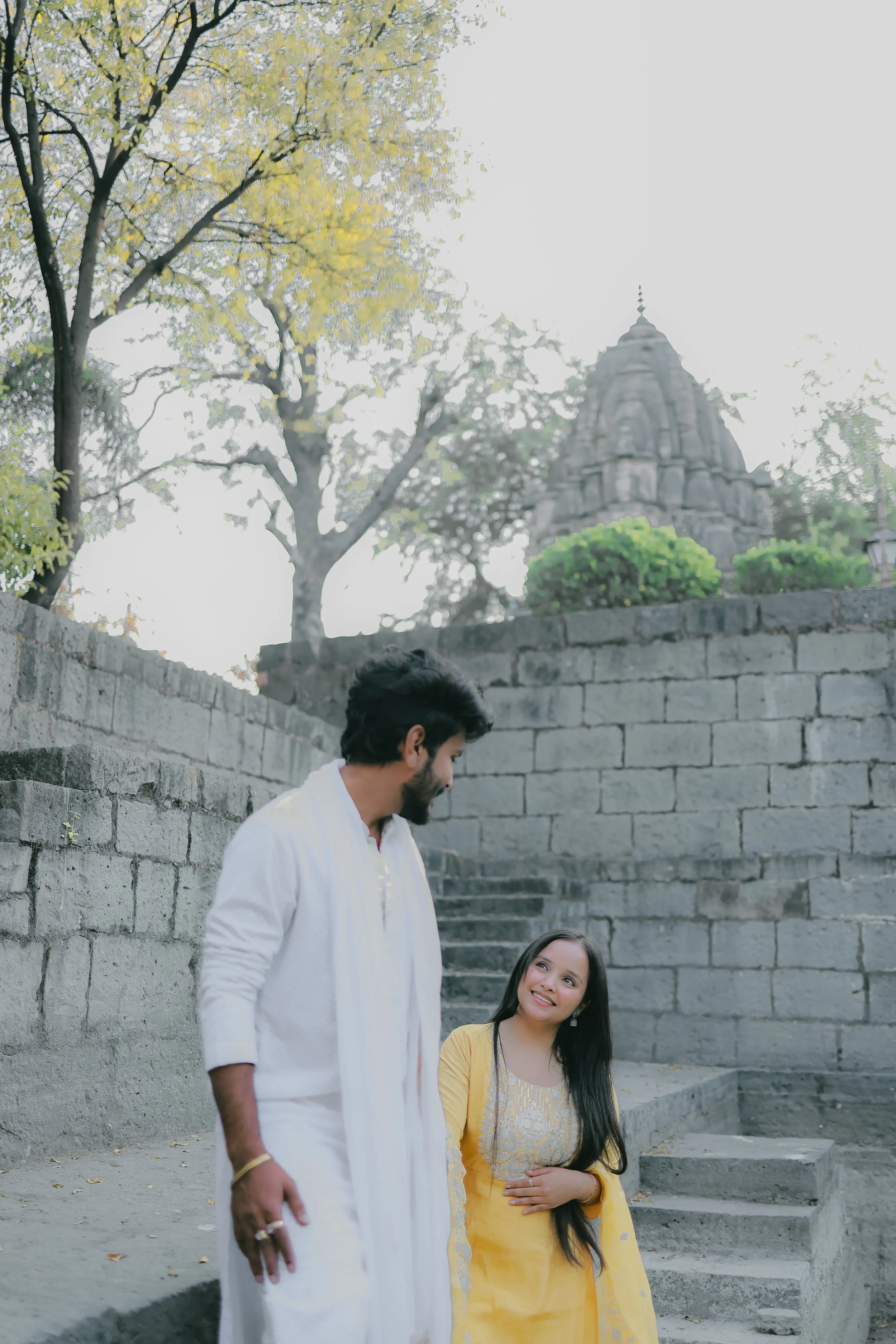 A man in a white traditional outfit and a woman in a yellow dress with embroidery are standing on stone steps outdoors, looking at each other. There is a stone wall behind them with steps leading up to a temple, and trees with yellow and green leaves