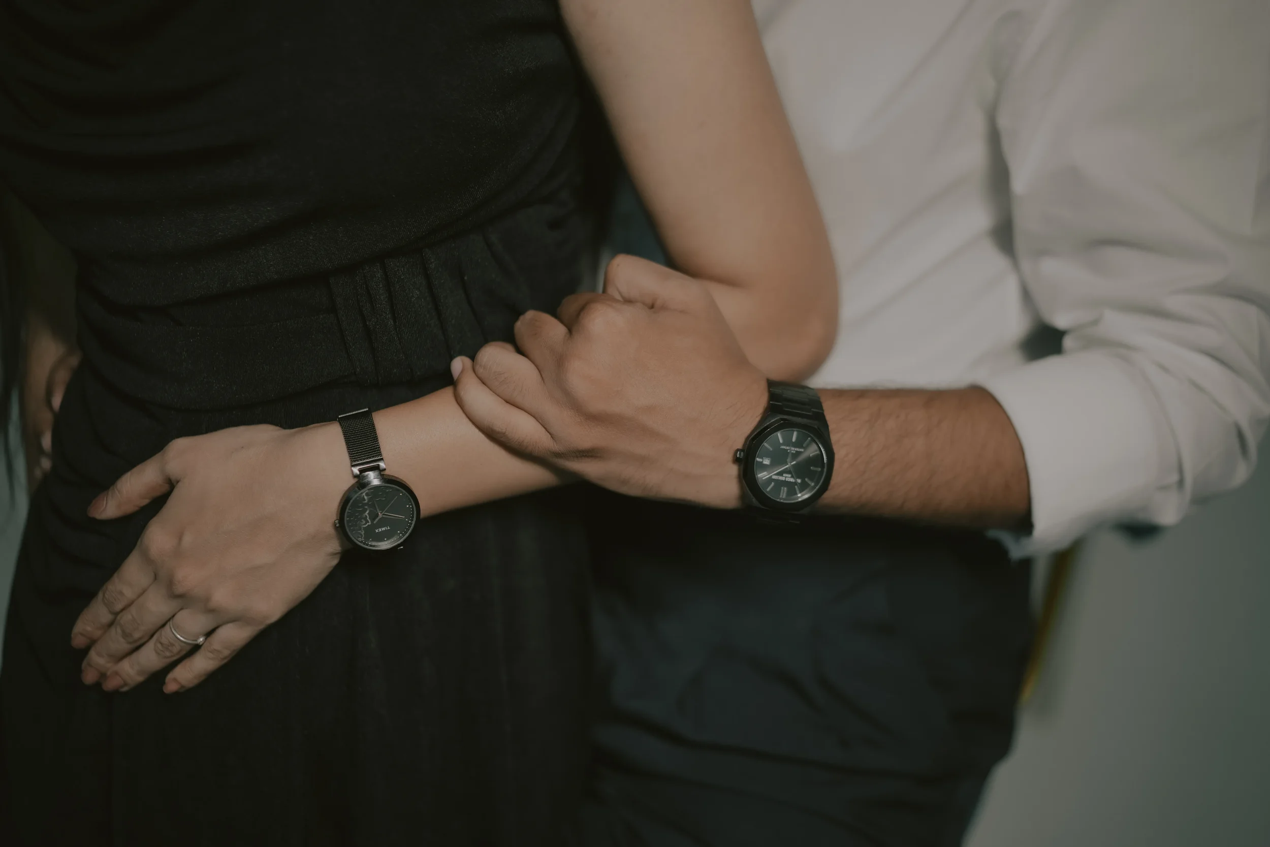 A person wearing a black wristwatch with silver accents and a black face, holding hands with another person who is wearing a similar watch, while sitting or lying on a bed with white sheets.