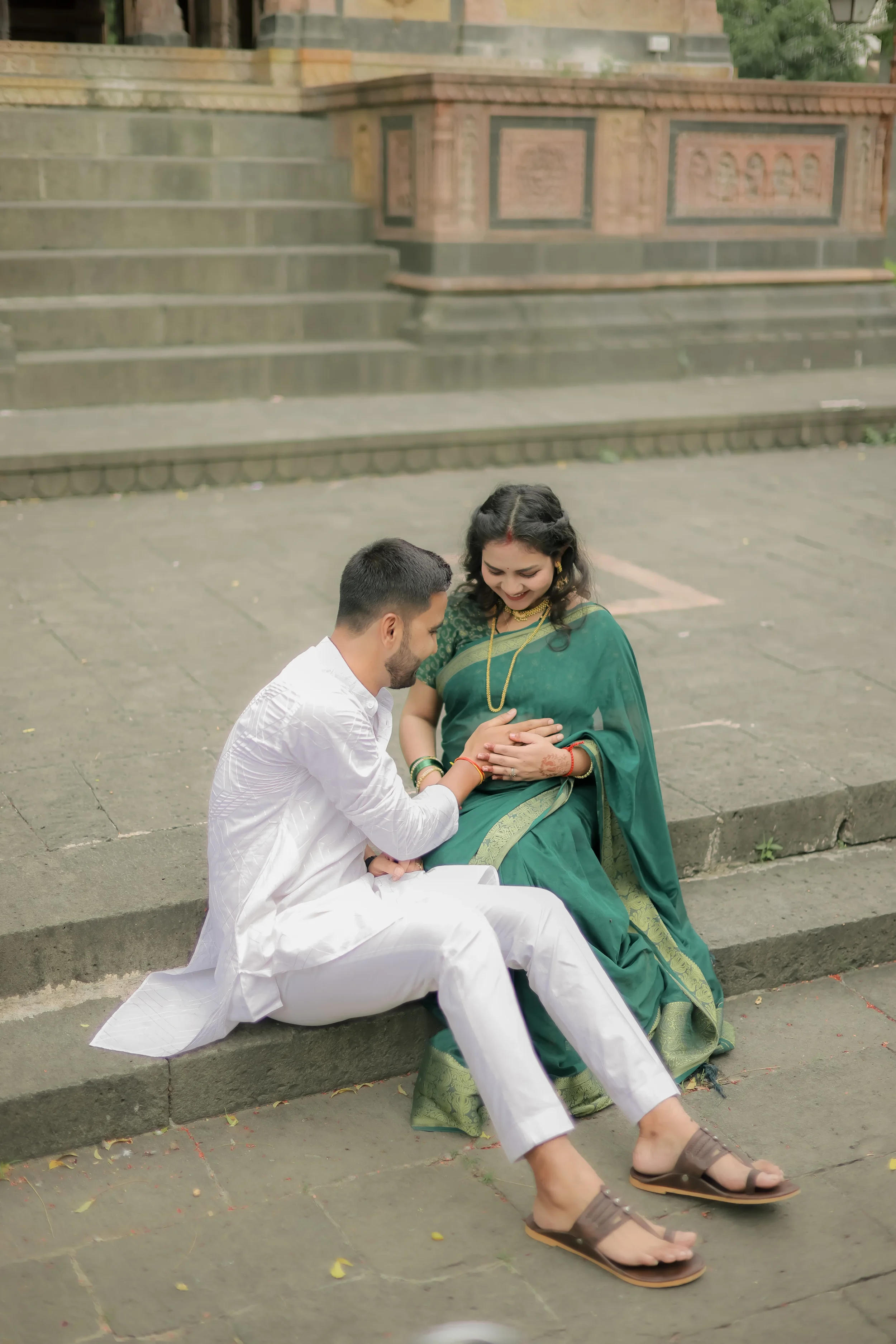 A couple of people sitting on a stone step in front of a historical building, one man dressed in white traditional attire and a woman in a green sari, both smiling.