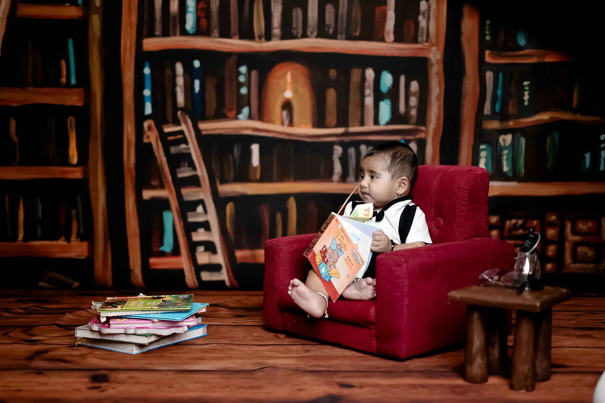 A young child with short dark hair sitting on a red armchair, holding a colorful children's book, with a pile of books on the wooden floor in front of him. A dark themed bookshelf mural decorates the background.