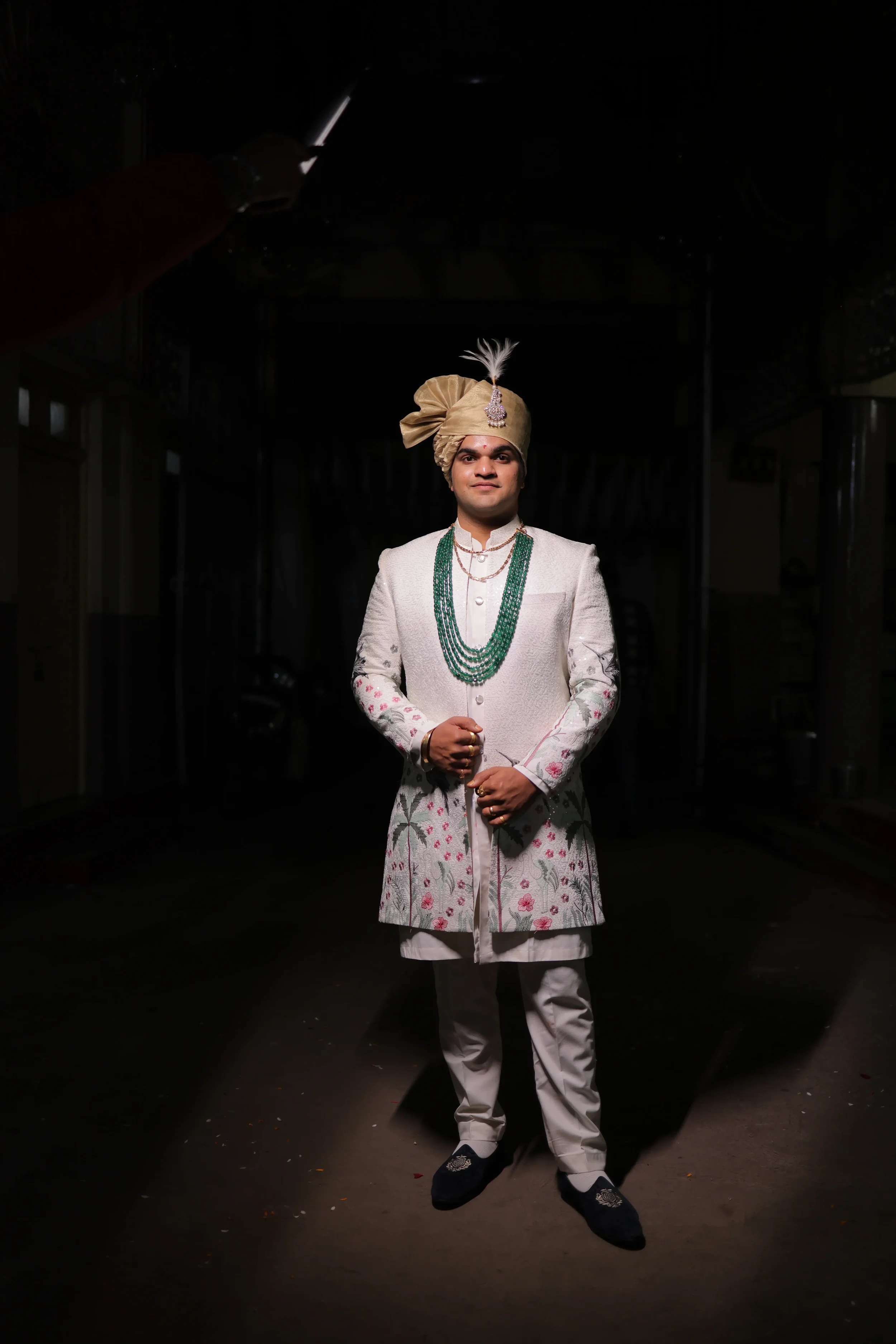 A man dressed in traditional Indian wedding attire standing with hands clasped in front, wearing a beige turban with a feather, a cream-colored sherwani with intricate embroidery, green beaded necklaces, white pants, and dark shoes.