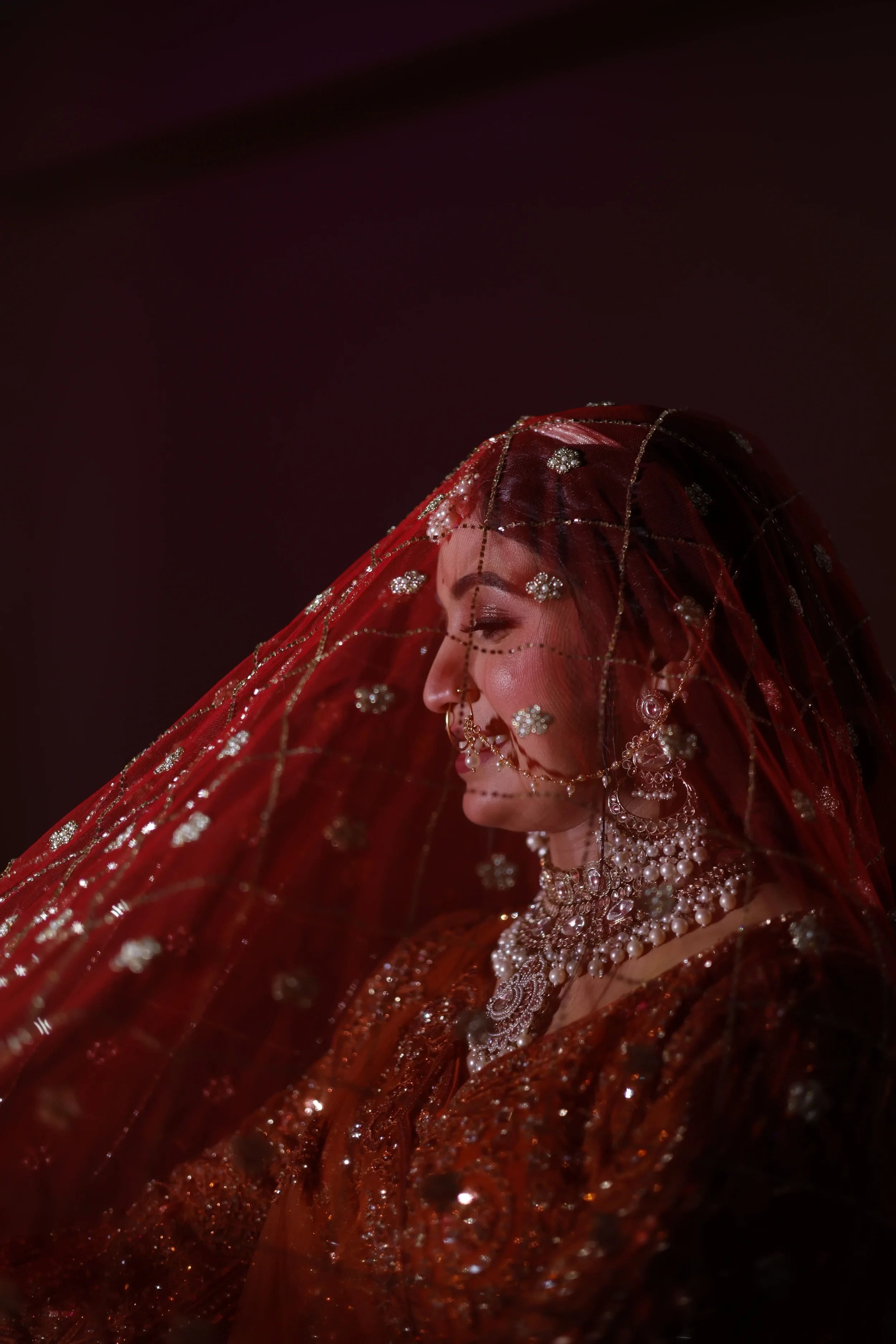 A woman is dressed in traditional South Asian bridal attire, wearing a red and gold bridal saree or lehenga with intricate beadwork and embroidery. She is adorned with elaborate jewelry, including a multi-layered pearl and gemstone necklace, matching earrings, and a decorative headpiece (maang tikka). Her face is partially visible, with makeup highlighting her features, and she is positioned against a dark background.