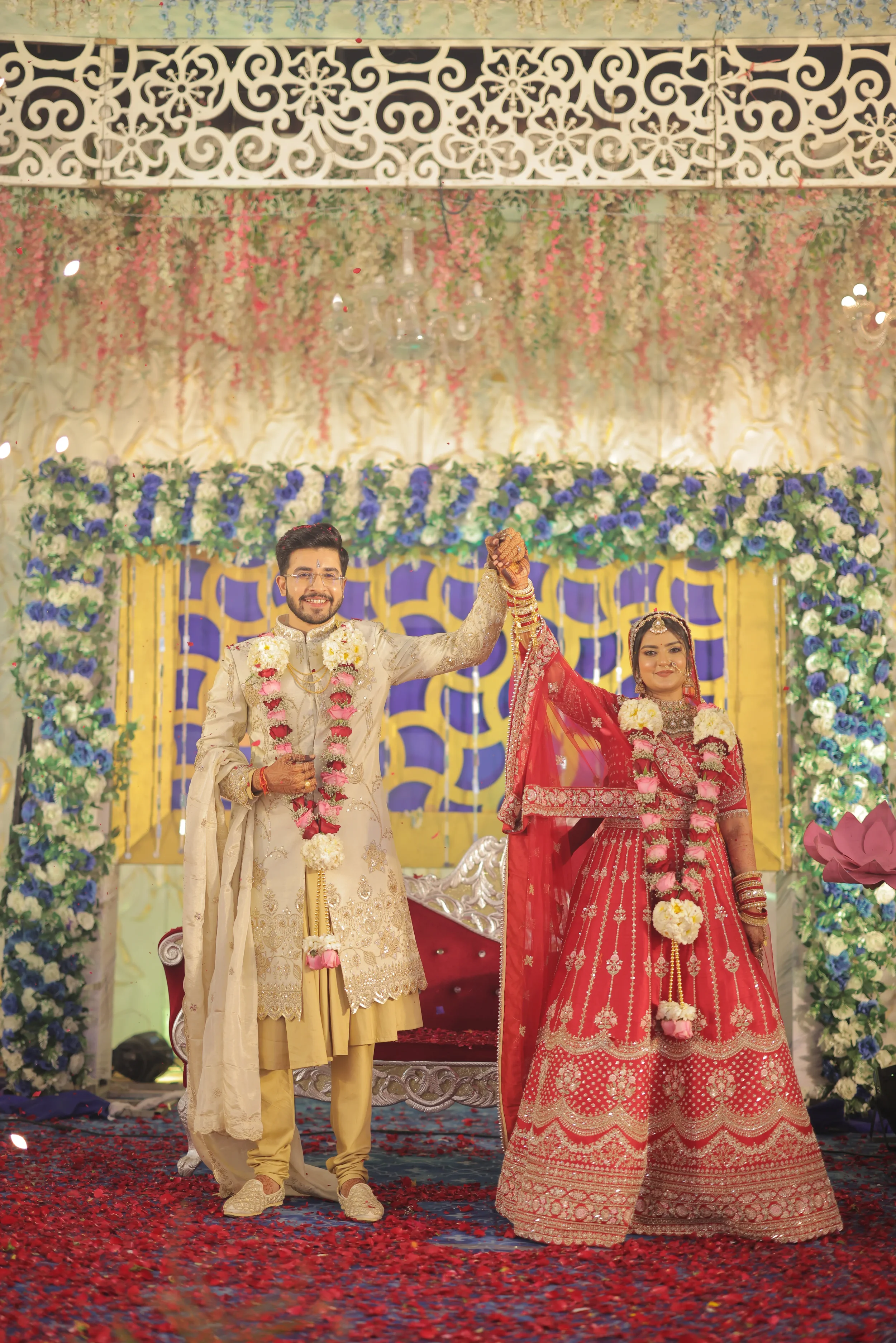 A newlywed Indian couple dressed in traditional wedding attire, holding hands on stage during their wedding celebration, surrounded by floral decorations and a patterned backdrop.