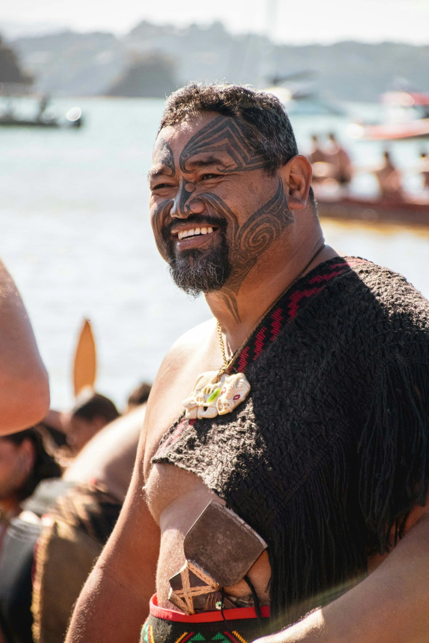 A man with traditional tattoos on his face and arm, wearing a necklace made of shells, participating in a cultural ceremony on the beach with boats in the background.