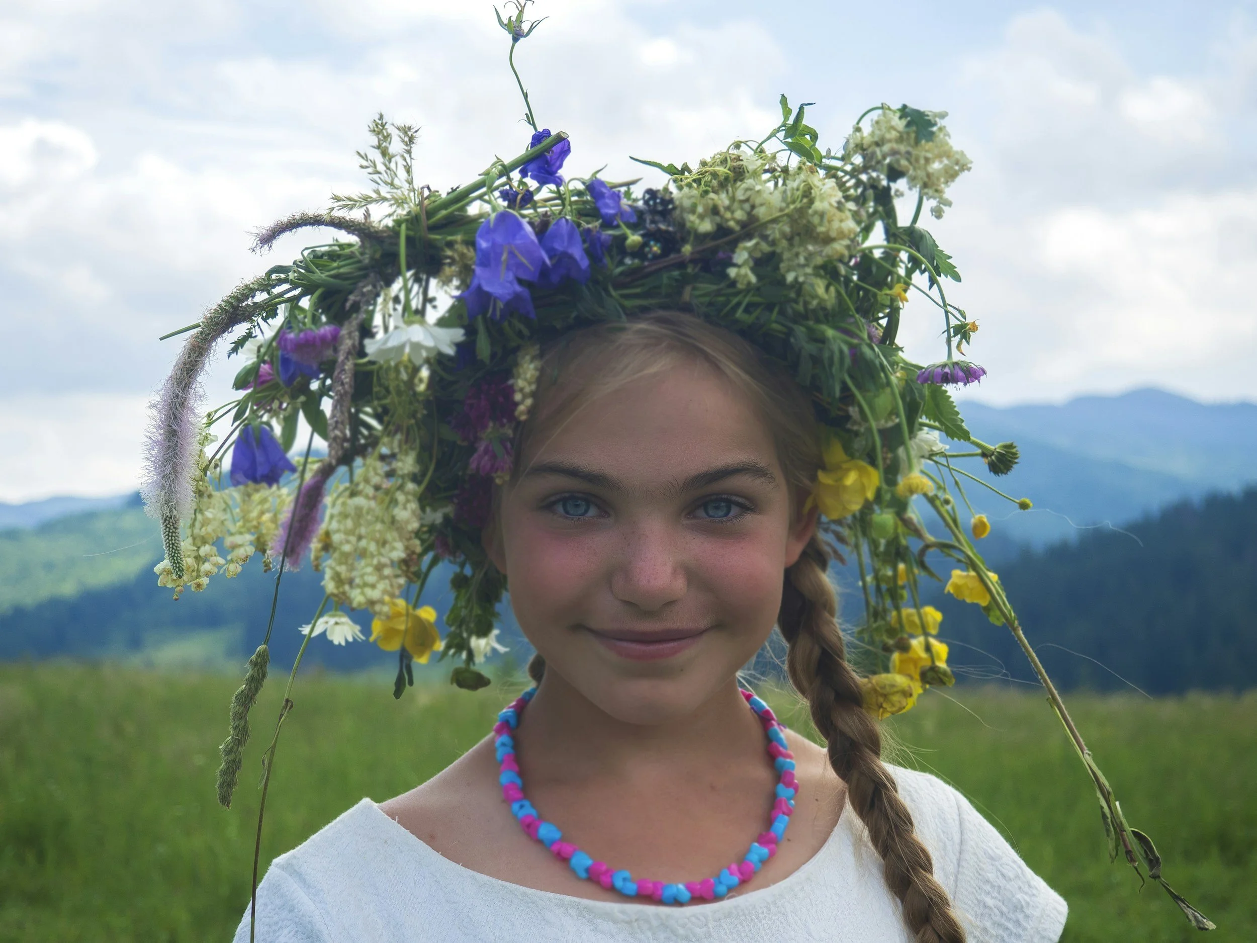 A young girl with braided hair wearing a white shirt, colorful beaded necklace, and a crown made of various flowers and greenery, standing outdoors with a mountain and cloudy sky in the background.