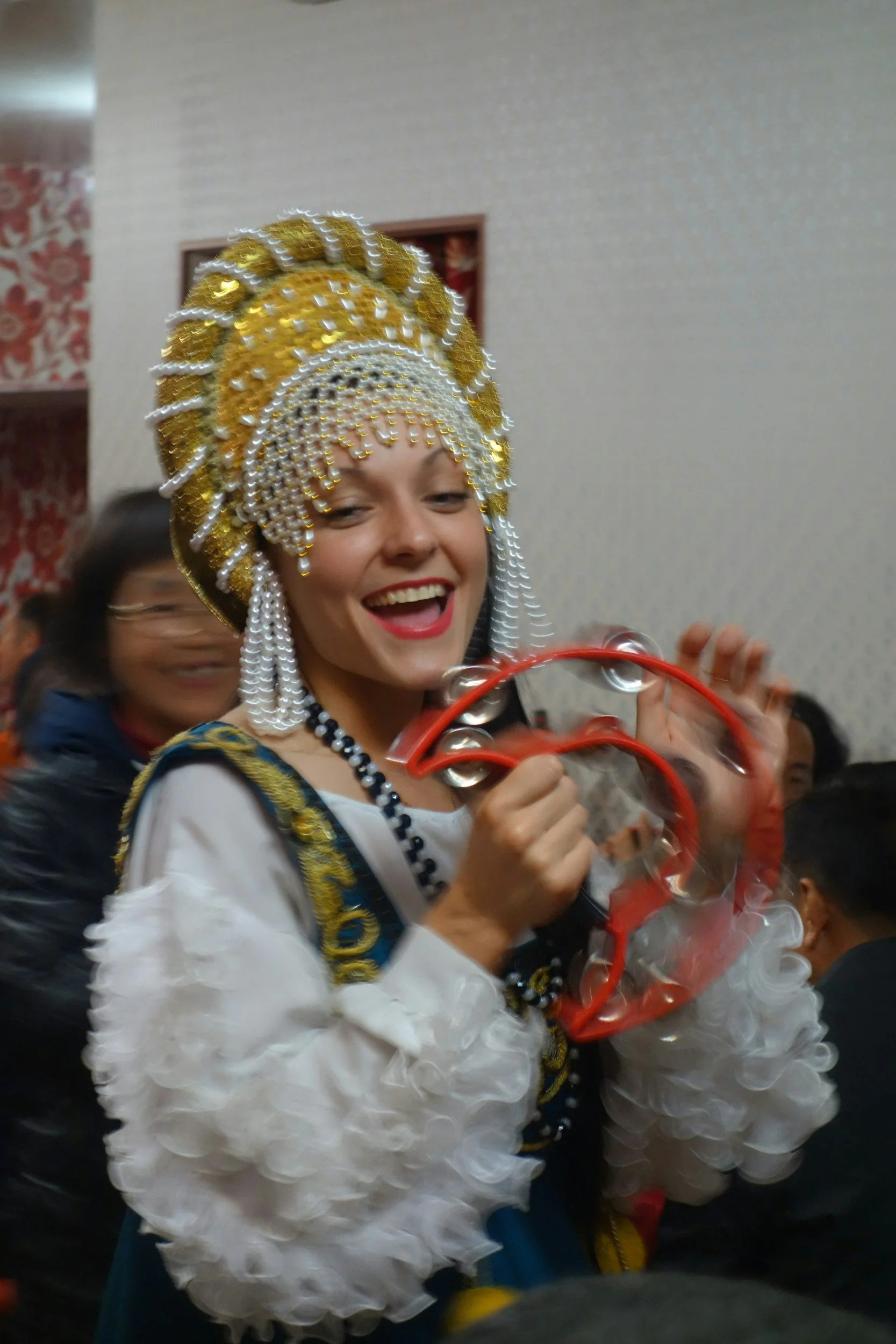 Woman in elaborate traditional costume with gold and white headdress, holding red tambourine, smiling at celebration.
