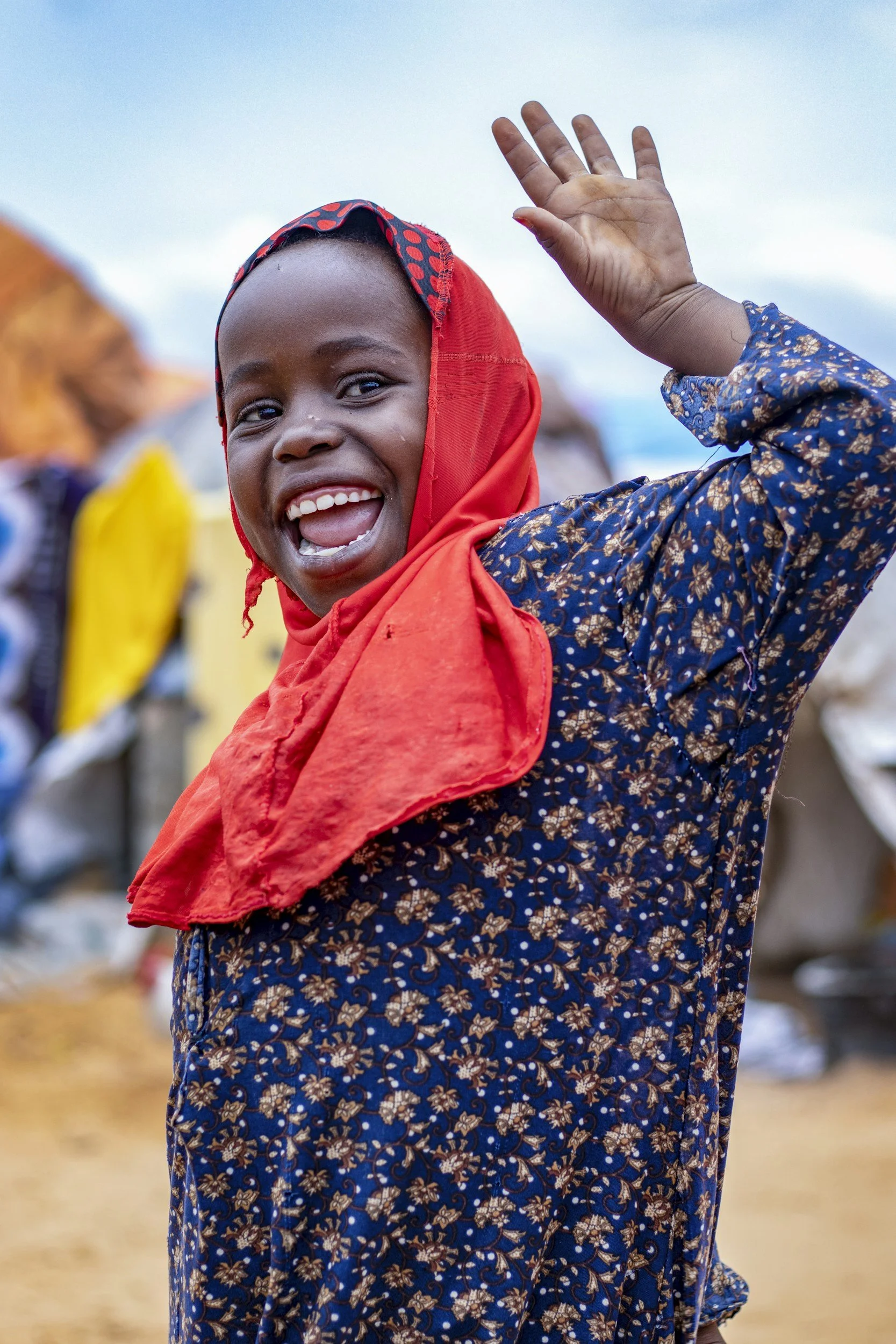 A joyful young girl wearing a blue patterned dress with a red shawl waving and smiling at the camera, outdoors with a bright sky behind her.