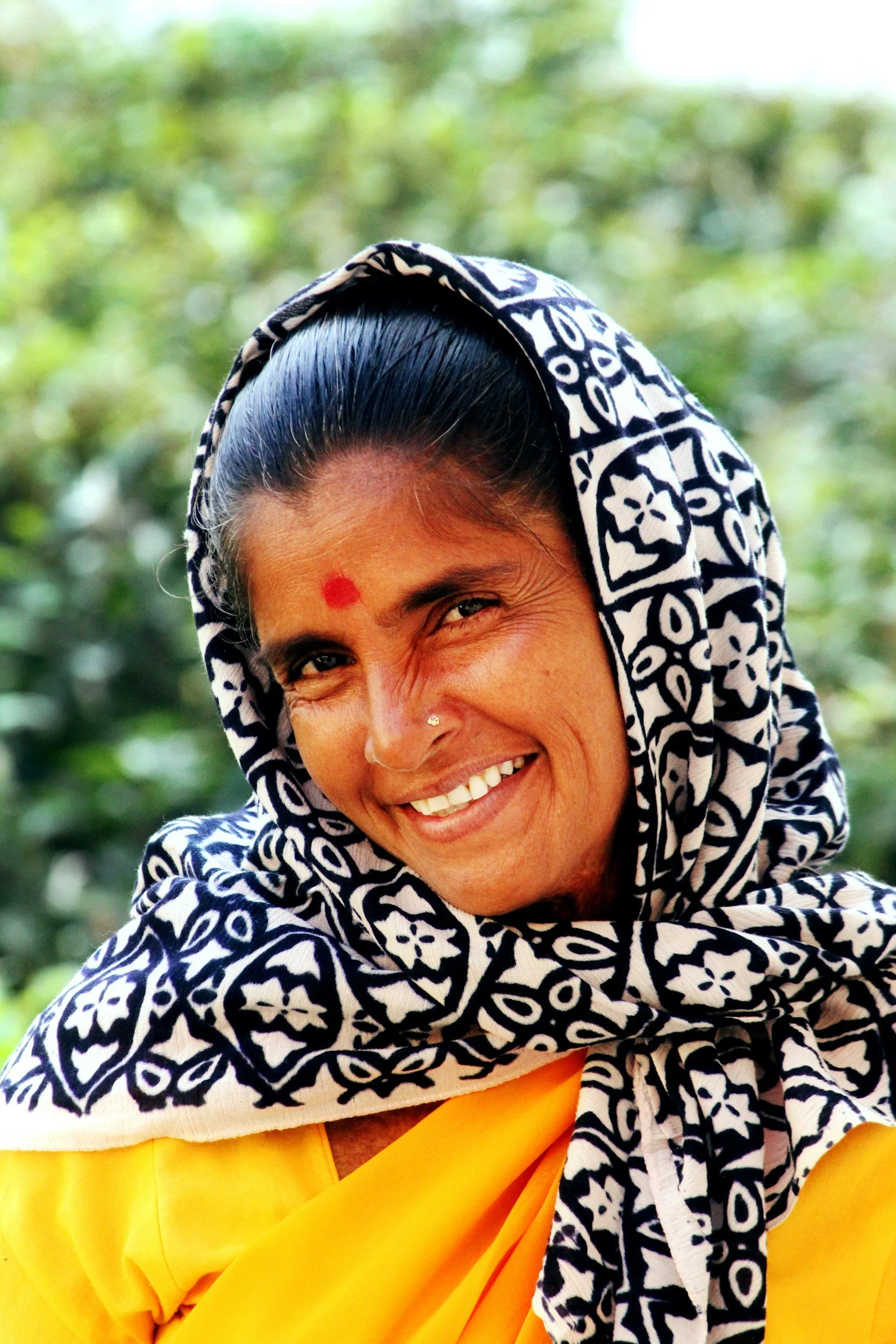 Close-up of a smiling woman with a red bindi on her forehead, wearing a black and white patterned headscarf and yellow clothing, outdoors with green foliage in the background.