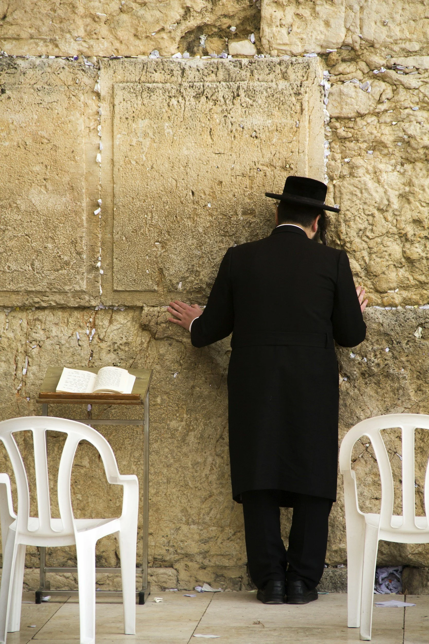 A man wearing traditional Jewish attire, including a black hat and suit, is praying at the Western Wall, a sacred Jewish site in Jerusalem. He is facing the wall with his hands resting on it, and there are white plastic chairs and a small table with an open prayer book nearby.