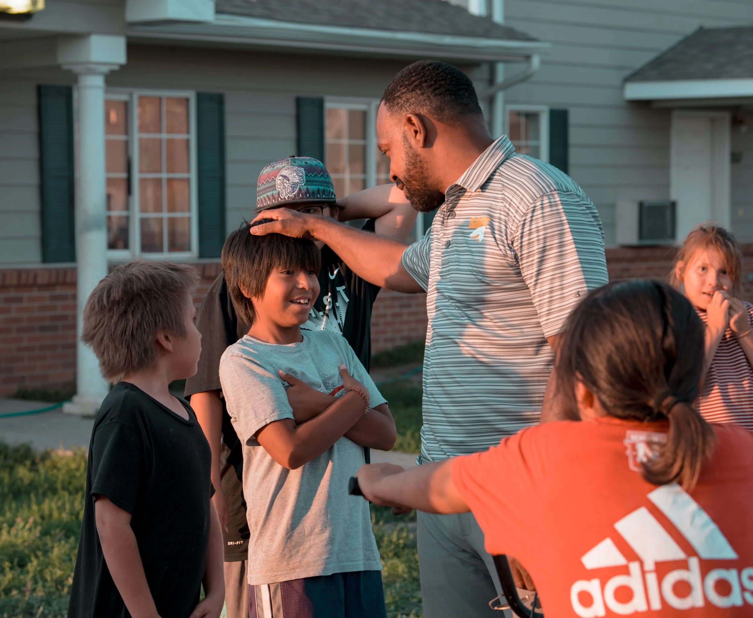 A man with a beard in a striped shirt is speaking to a group of children outdoors in front of a house, during sunset.