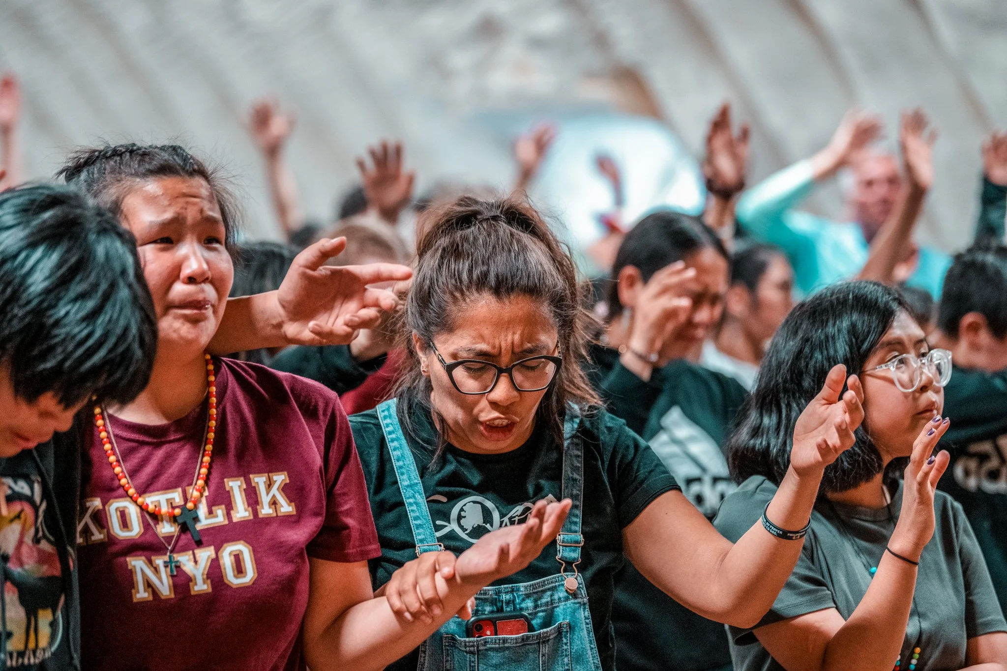 A group of people with diverse ethnicities appear to be praying or meditating, with some eyes closed and hands raised or clasped, in an indoor setting.