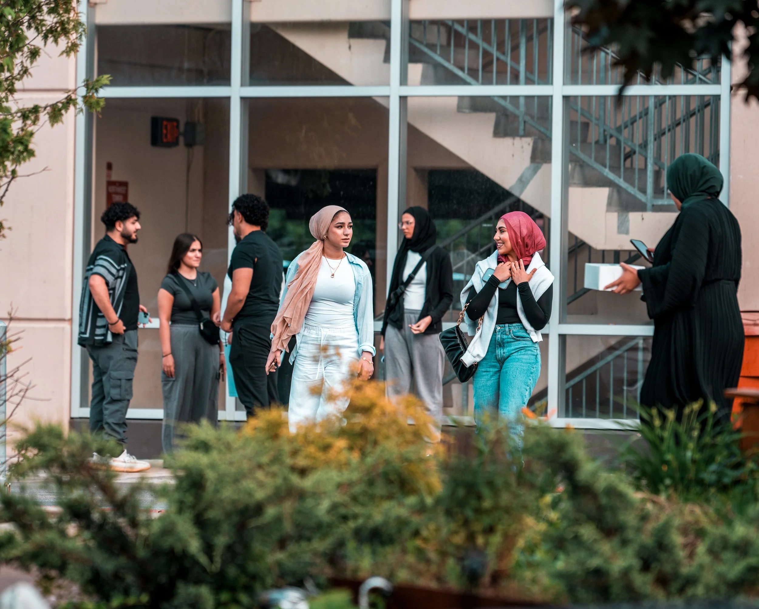Group of diverse young adults standing and talking outside a building with glass windows and a staircase, some with phones in hand.