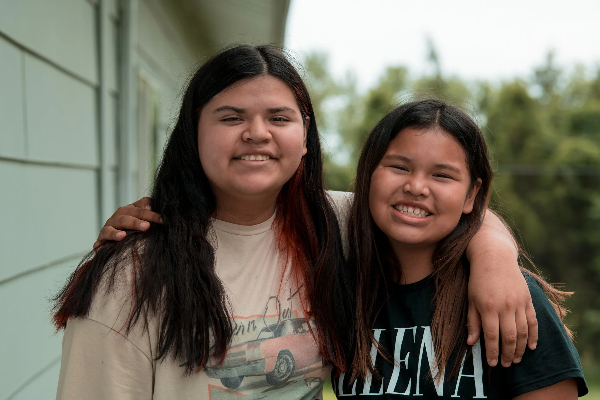 Two young women with long dark hair and tan skin smiling and hugging outside in front of a light-colored house and trees.
