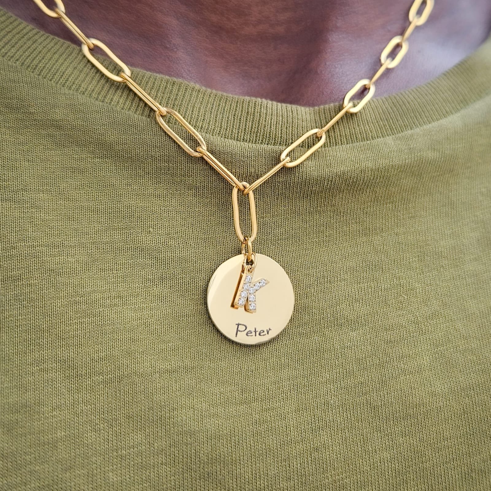 Close-up of a person wearing a gold chain necklace with a circular pendant that has the letter 'K' decorated with diamonds, as well as the name 'Peter' engraved on the pendant.