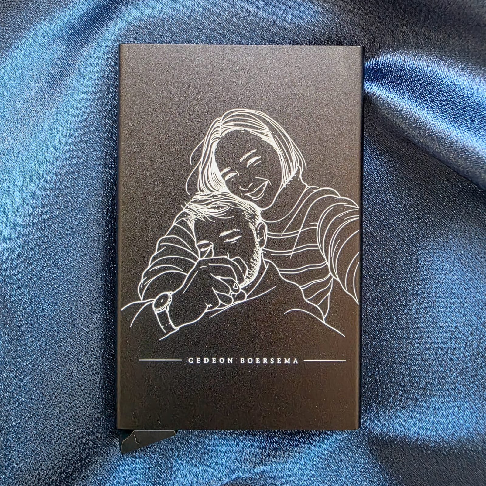 Black credit card holder engraved with white line art of a smiling woman holding a man with his name at the bottom.