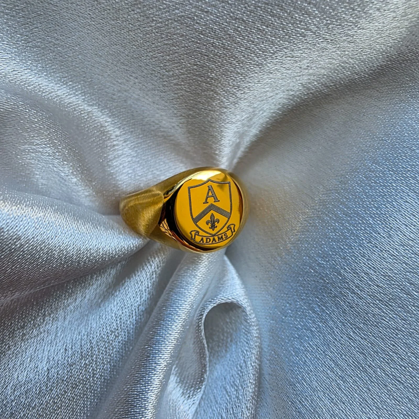 A golden signet ring with a shield-shaped emblem featuring the letter A, a fleur-de-lis, and the word 'Adams' engraved on it, resting on shiny silver fabric.