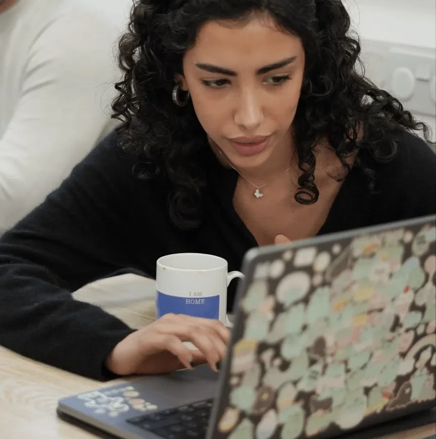A woman with curly black hair wearing a black top, silver hoop earrings, and a necklace with a four-leaf clover pendant is sitting at a table, looking at a laptop with a colorful, patterned cover. She has a white mug with blue accents on the table in front of her.