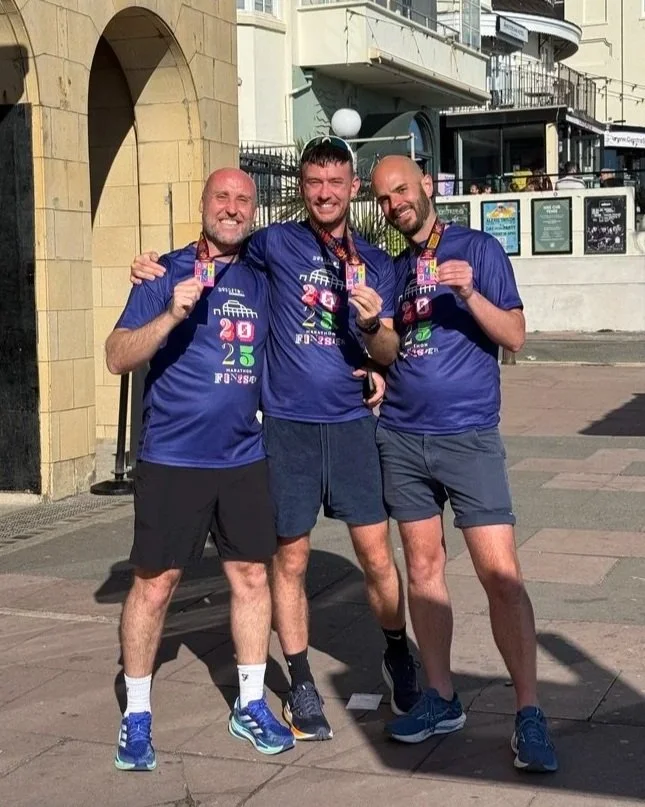 Three men celebrating with medals in front of a building after running the Brighton Marathon, all wearing blue shirts and shorts, smiling.