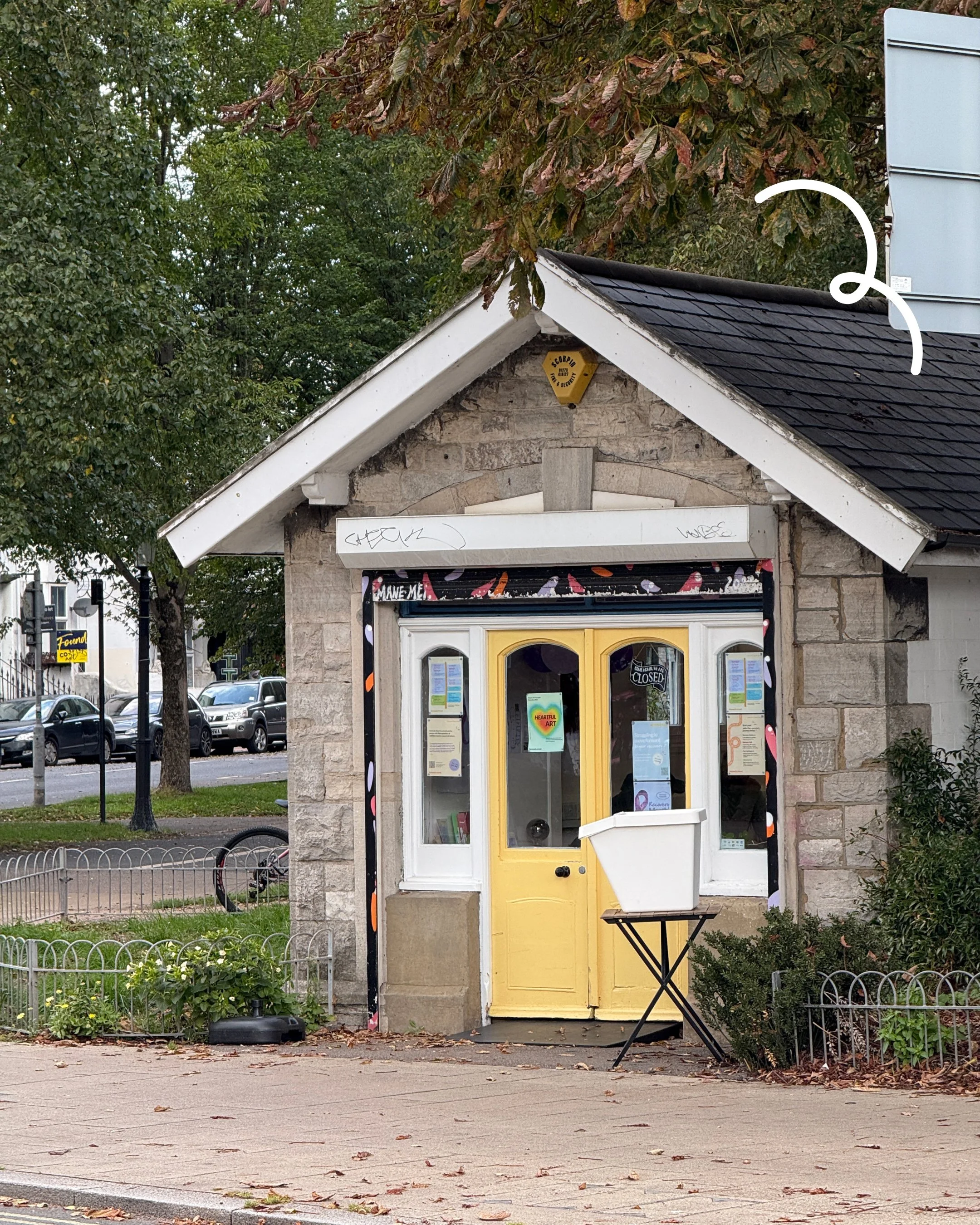 Small building with a stone facade and a yellow door, featuring a white sign and colorful decorations around the door frame, situated along a sidewalk with trees and parked cars in the background.