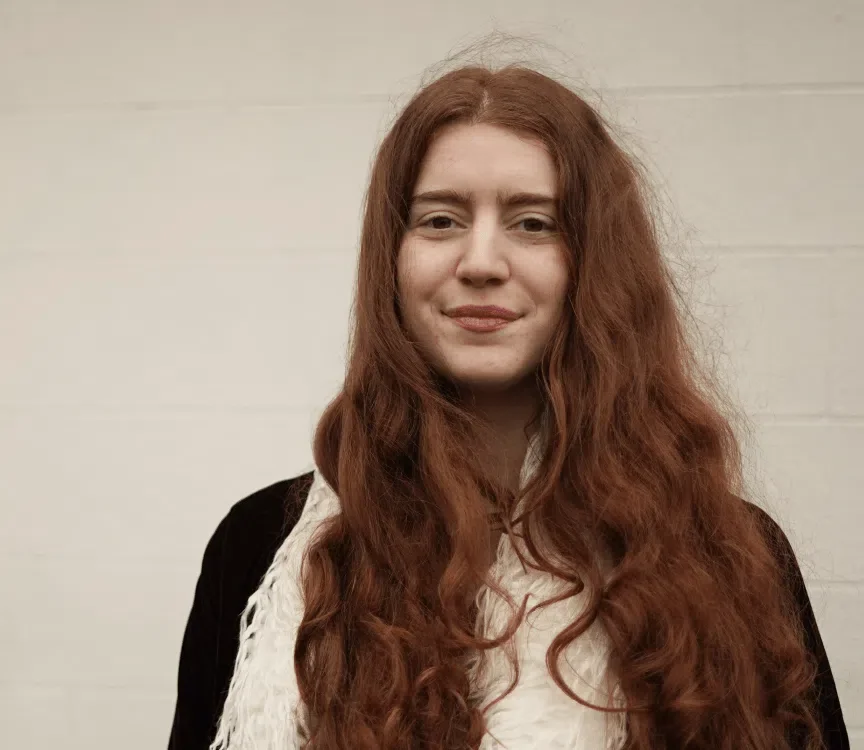 A woman with long, wavy red hair, wearing a black jacket and a white scarf, standing against a light-colored wall.