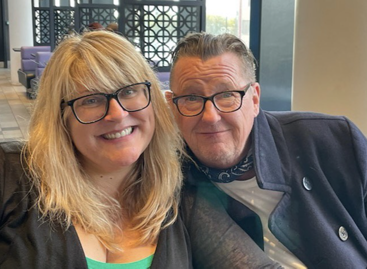 A smiling woman with blonde hair and glasses, and a man with glasses and short hair, both in casual attire, sitting close together in a modern indoor setting.