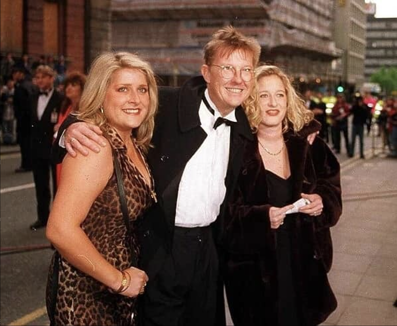 Three people, two women and one man, dressed in formal attire, standing together outdoors at night during a public event or celebration.