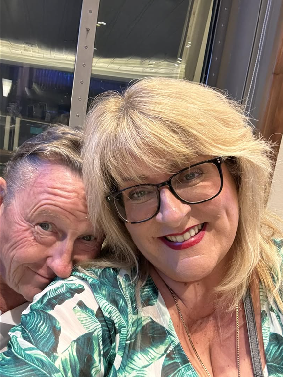 A woman with blonde hair, glasses, and red lipstick smiling at the camera, next to an older man with gray hair, leaning close to her. They appear to be in an indoor setting with a window or glass behind them.