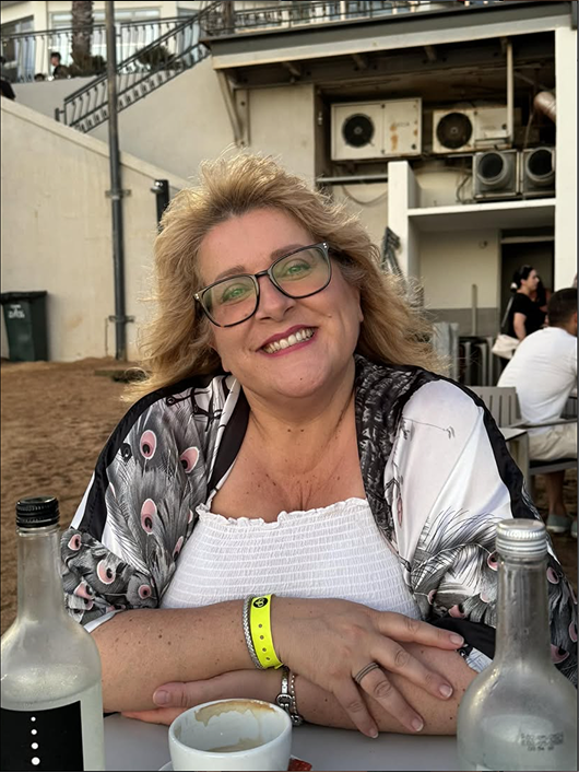 A smiling woman with blonde hair and glasses sitting outdoors at a table with bottles and a cup, with other people and building in the background.