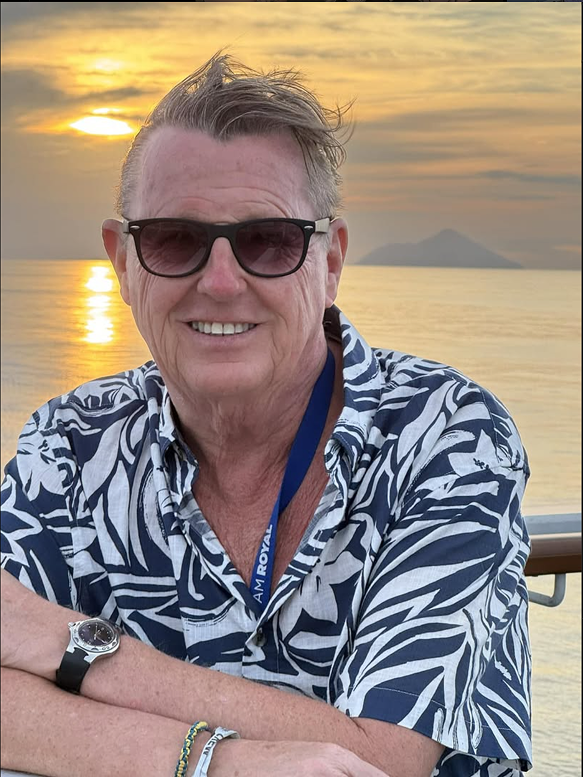 A smiling man wearing sunglasses and a patterned short-sleeve shirt, enjoying a sunset by the water with an island in the background.