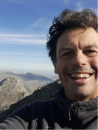 Man taking a selfie outdoors on a mountain with a view of rocky peaks and a blue sky.