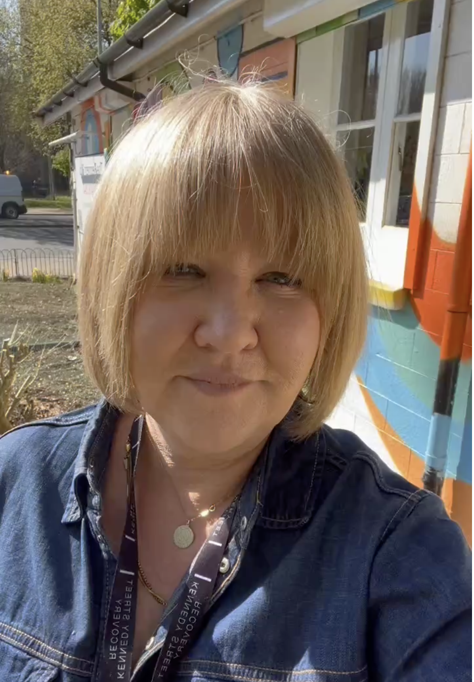 A woman with blonde hair taking a selfie outdoors in front of a colorful mural on a building. She is wearing a denim jacket and a lanyard with text.