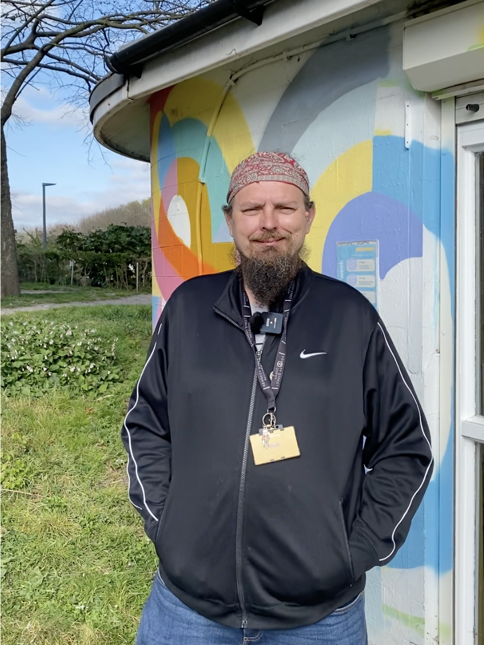 A man with a beard and mustache wearing a red bandana, black Nike jacket, and jeans, standing outdoors in front of a colorful mural on a building, with a grassy area and trees in the background.