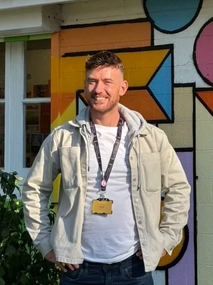A young man with short hair and a beard, smiling, standing outside in front of a colorful graffiti wall, wearing a light beige jacket, white t-shirt, and a camera around his neck.