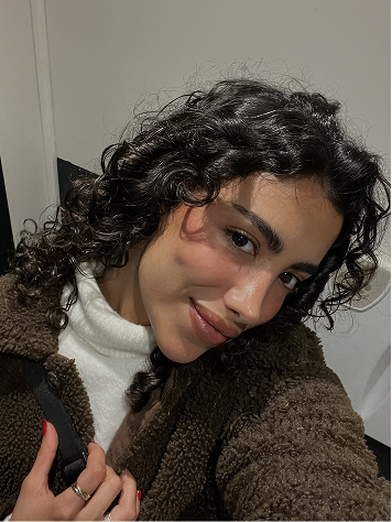 A woman with curly dark hair taking a selfie, wearing a white turtleneck and a brown fleece jacket, with a neutral background.